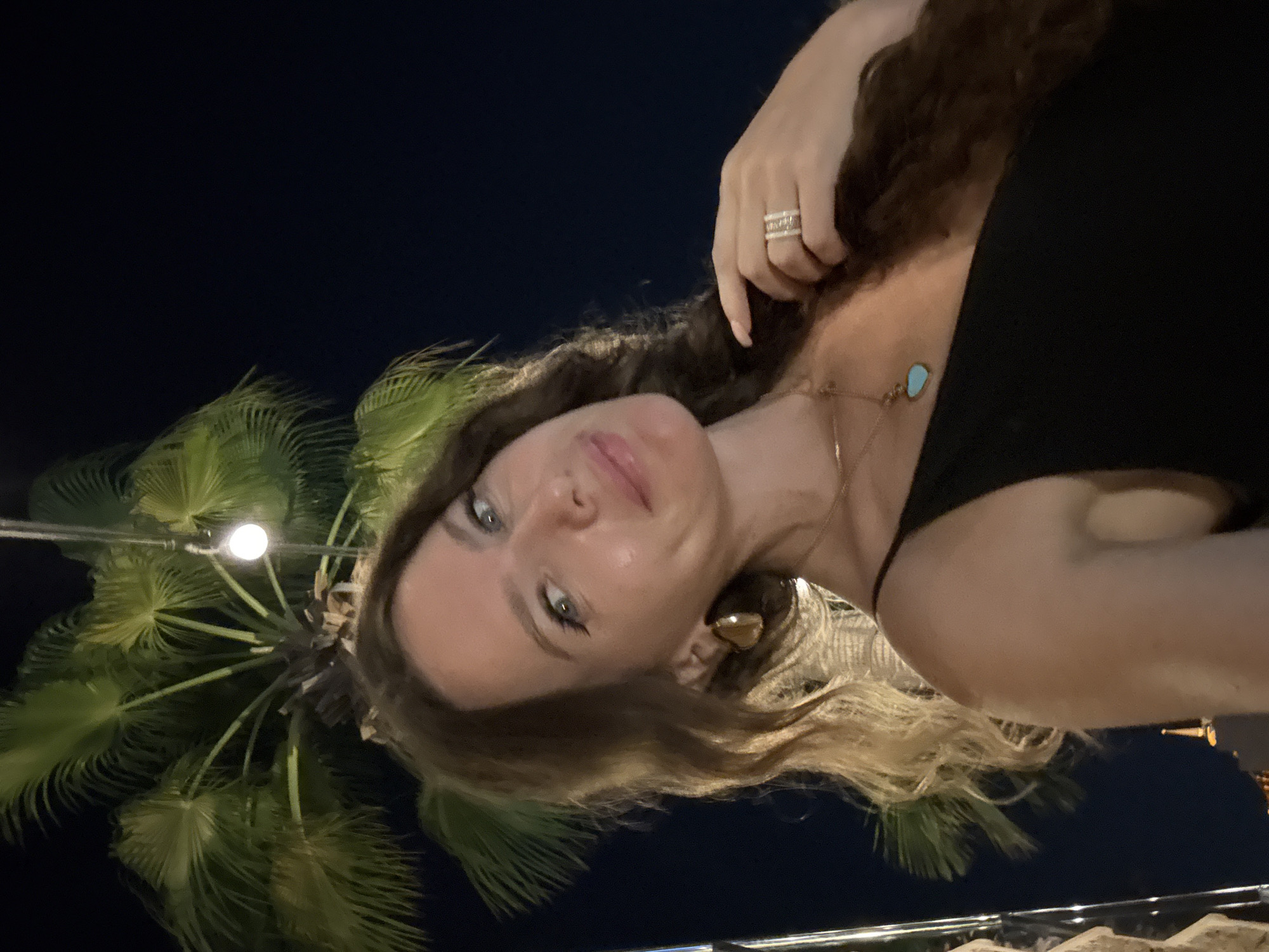 A woman is photographed at night, possibly in Dubai, United Arab Emirates, against a backdrop of a palm tree and a dark sky. She has long, wavy brown hair and is wearing a black dress, a gold teardrop earring, and rings on her right hand. A necklace with a blue, heart-shaped pendant is visible. Her right hand is touching her hair near her face. The lighting suggests an outdoor setting, possibly a restaurant or event space.