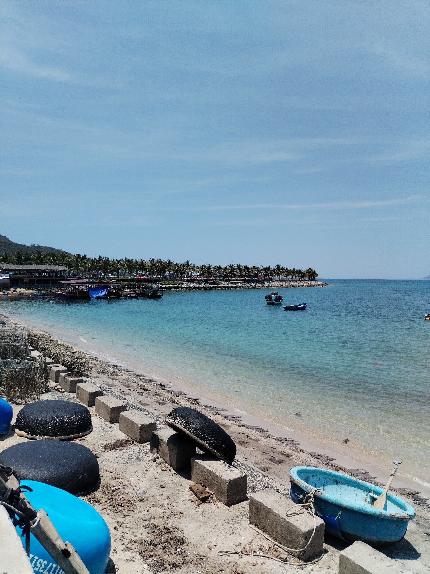 The image captures a coastal scene in Bac Nha Trang Ward, Vietnam. The foreground consists of a sandy beach littered with debris and concrete blocks, seemingly forming a rudimentary barrier. Several circular fishing boats, painted in black and blue, are scattered along the beach. A turquoise-colored boat sits partially on the beach, complete with a wooden oar inside.
Beyond the beach, the clear, turquoise water of the sea stretches to the horizon, dotted with small fishing boats. In the distance, a coastline is visible, lined with buildings and palm trees, possibly indicating a populated area. The sky is mostly clear, with scattered clouds. The overall scene suggests a working beach environment with a mix of natural beauty and human activity.