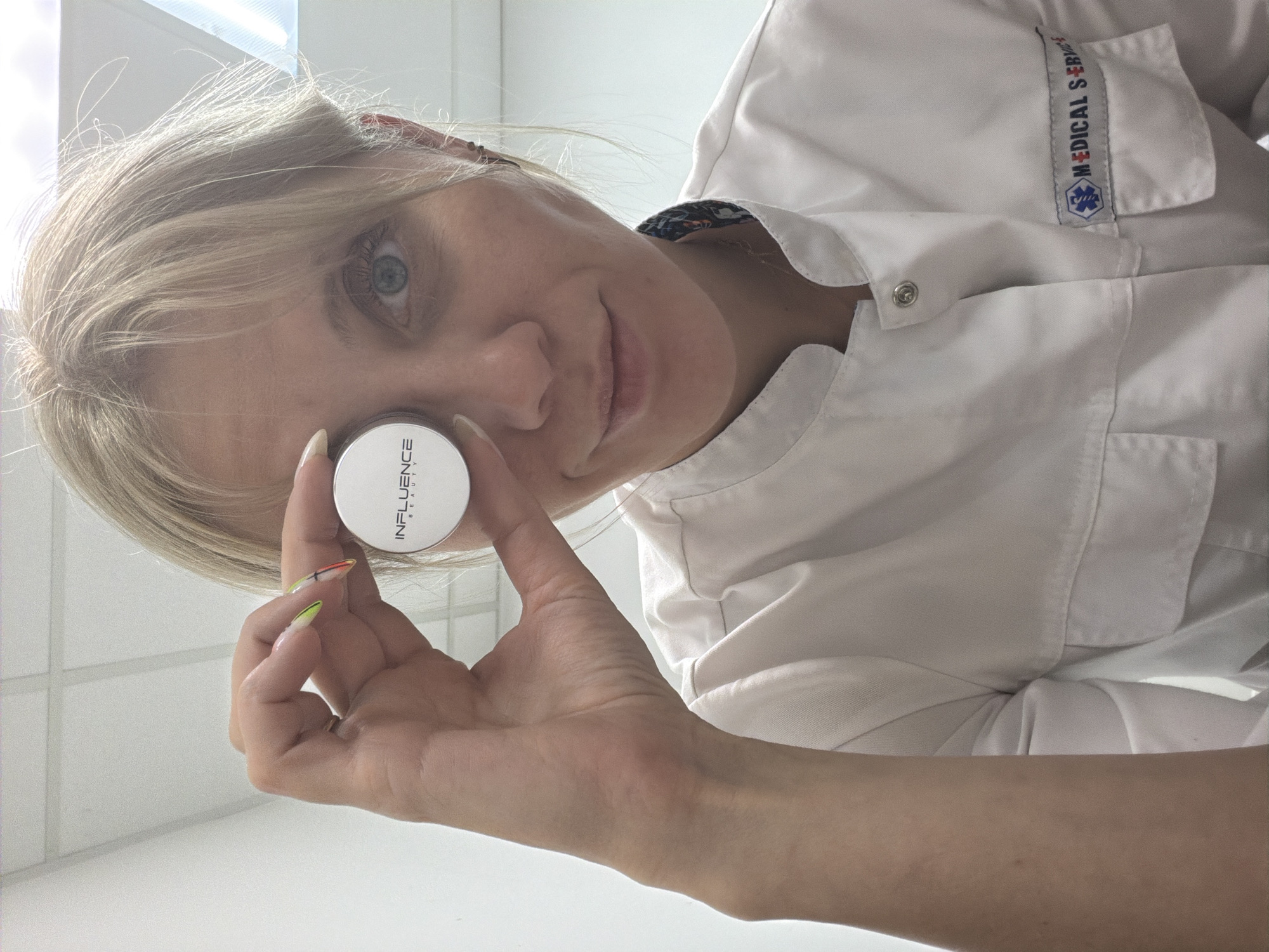 A Caucasian woman with blonde hair and blue eyes, wearing a white "MEDICAL SERVICE" shirt, is holding a white jar labeled "INFLUENCE BEAUTY" near her right eye. The scene appears to be indoors. The location is the city of Chaikovsky, Russia.