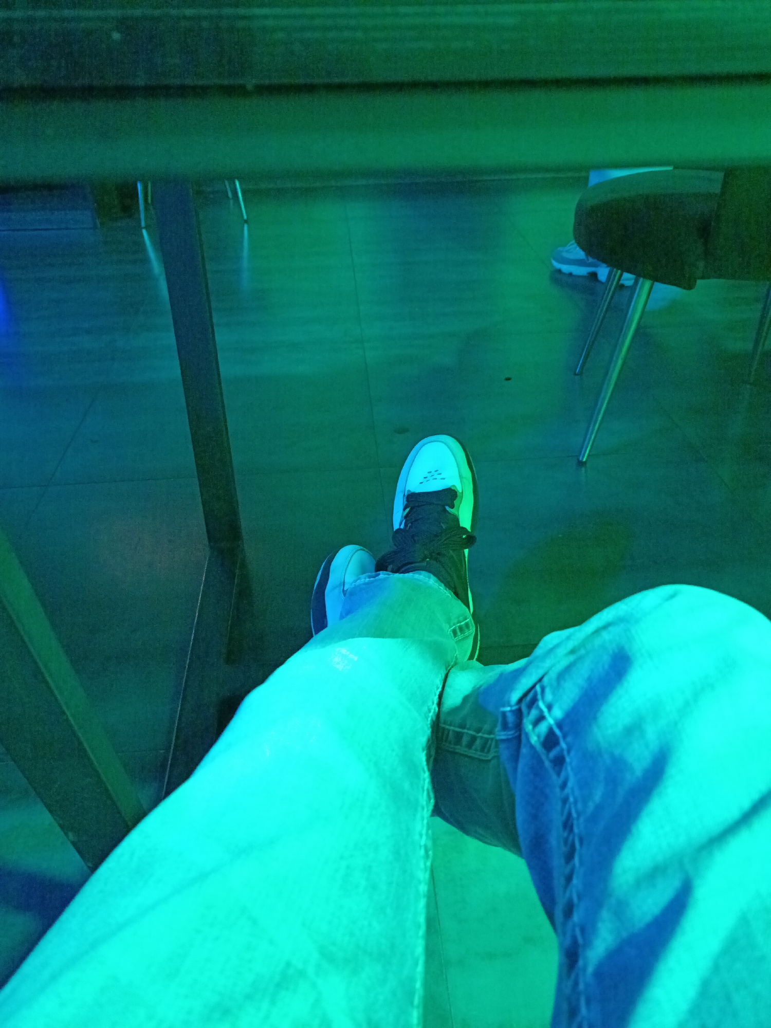 This is a low-angle, medium shot of a person's legs and feet in a dimly lit room. The person is wearing light-wash denim jeans and white and black sneakers. The scene is illuminated by green and blue lighting, casting a cool-toned glow on the floor and surrounding objects. A wooden table is visible in the foreground, and a chair with metallic legs can be seen in the background. The floor appears to be tiled. The image focuses on the lower extremities of the individual, with the upper body and face not visible. The overall impression is of a casual, modern setting.