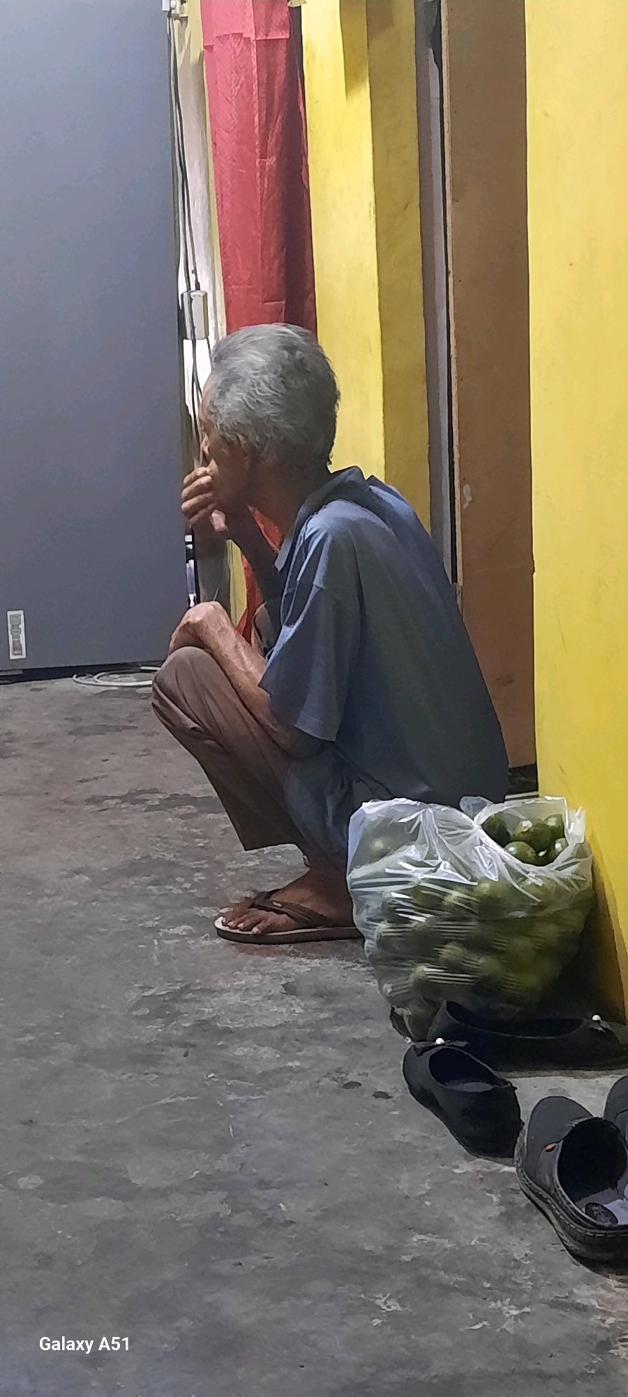 An elderly person with gray hair is squatting on a concrete floor, facing away from the camera and to the right. They are wearing a blue shirt and brown pants, with brown flip-flops on their feet. In front of them is a clear plastic bag filled with green citrus fruits, possibly limes or lemons. To the right of the bag, two pairs of black shoes are visible on the floor. Behind the person, there are colored cloths hanging, one red and one yellow, against a yellow wall. The overall environment appears to be an outdoor or semi-outdoor space. The image is tagged "Galaxy A51". The location is identified as Selopuro, Indonesia.
