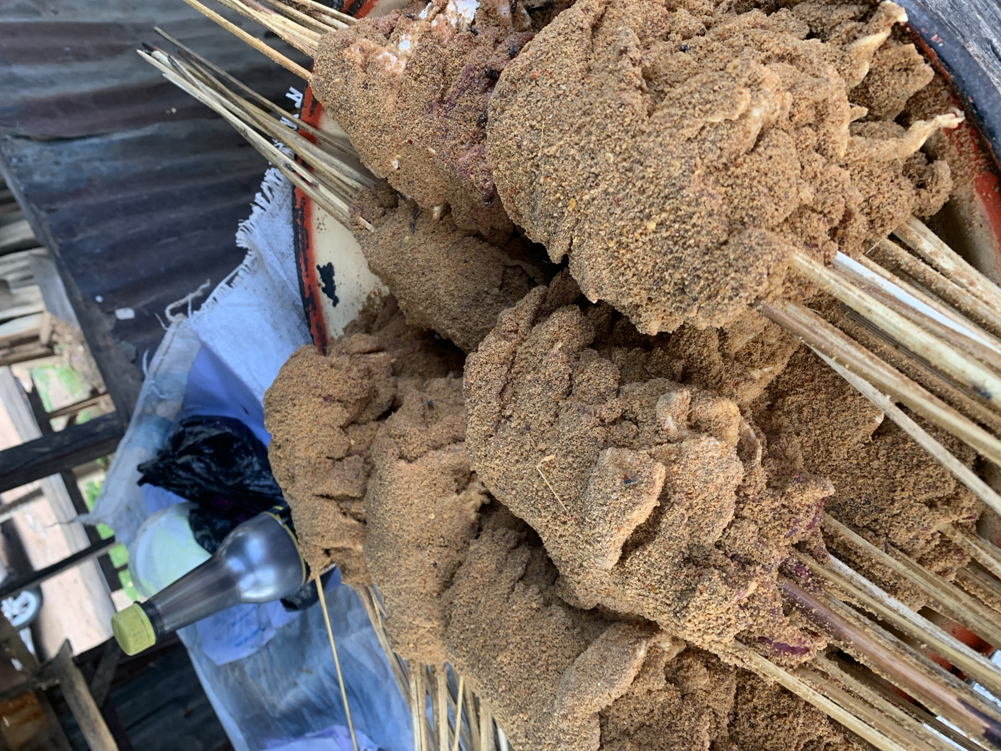 The image shows a close-up of skewered meat, heavily coated in a brown, grainy spice mixture. The skewers are piled together in a red-rimmed tray, resting on a light blue cloth covering a wooden surface. A silver-colored bottle with a yellow-green cap is visible, along with a black plastic bag. The environment appears to be outdoors, with a partial view of a structure with corrugated metal roofing. The location is New Stadium, Nigeria.