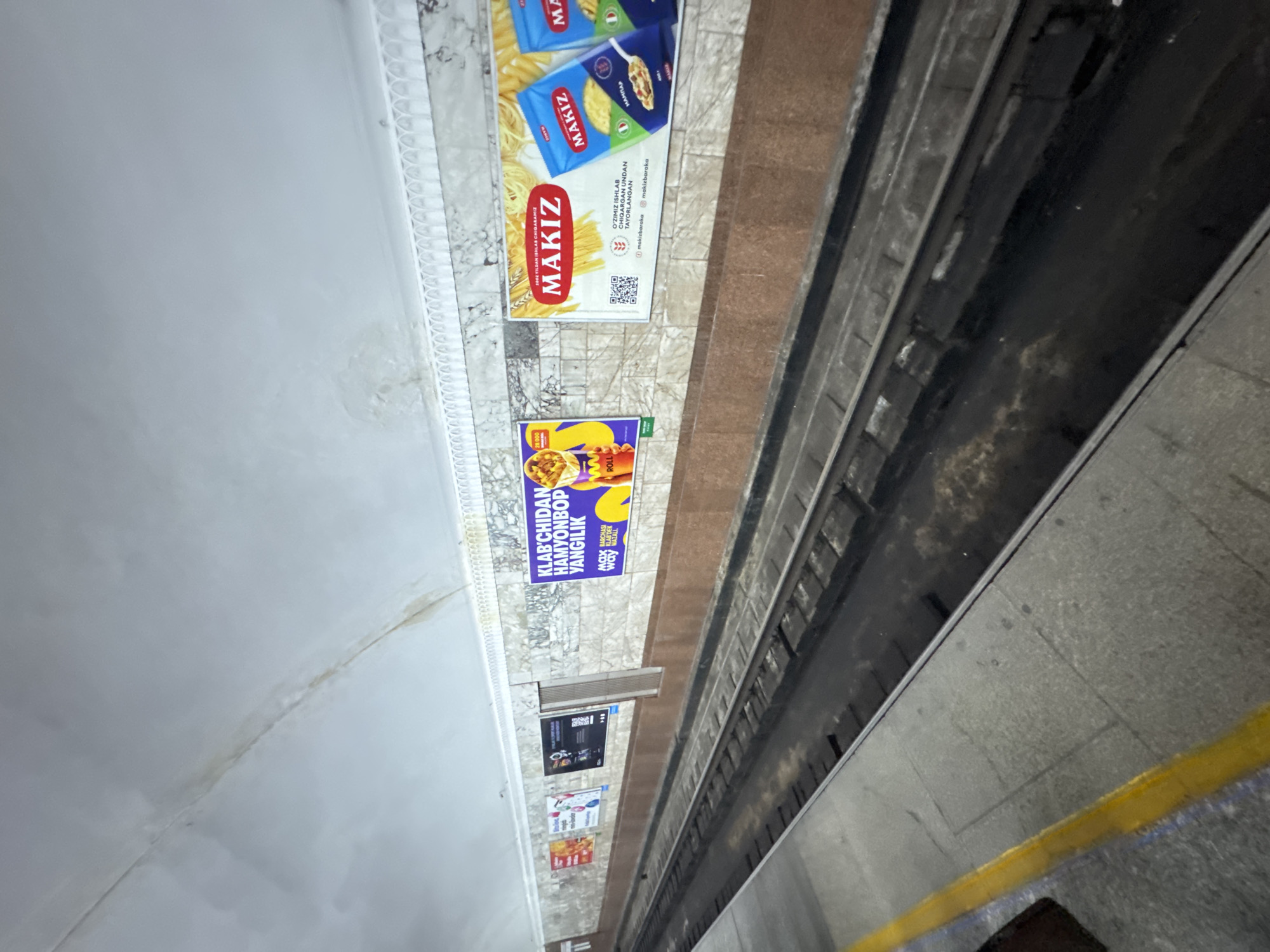 The image depicts a subway platform, likely in Tashkent, Uzbekistan. The platform is lined with advertisements, including one for "Makiz" pasta and another for "Roll" snacks. There is a yellow safety line along the edge of the platform. Subway tracks are visible. The image is taken at an angle, and lighting conditions suggest the location is indoors.