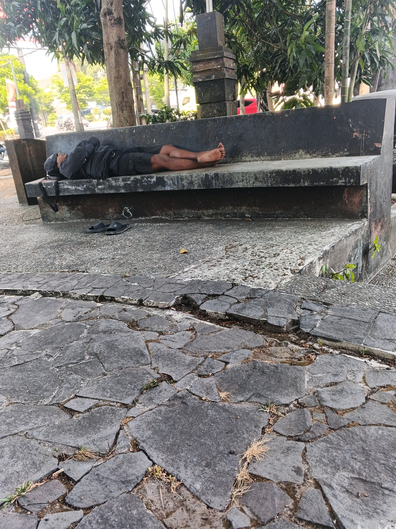 A person wearing a grey hoodie and black shorts is sleeping on a stone bench. The person is lying on their back with their head resting on their folded arms, and their feet are visible at the end of the bench. A pair of black sandals are placed on the ground in front of the bench. The bench is situated outdoors in what appears to be a park or public space with trees in the background. The ground in front of the bench is paved with cracked stone tiles. The scene is set in Ungaran, Indonesia.