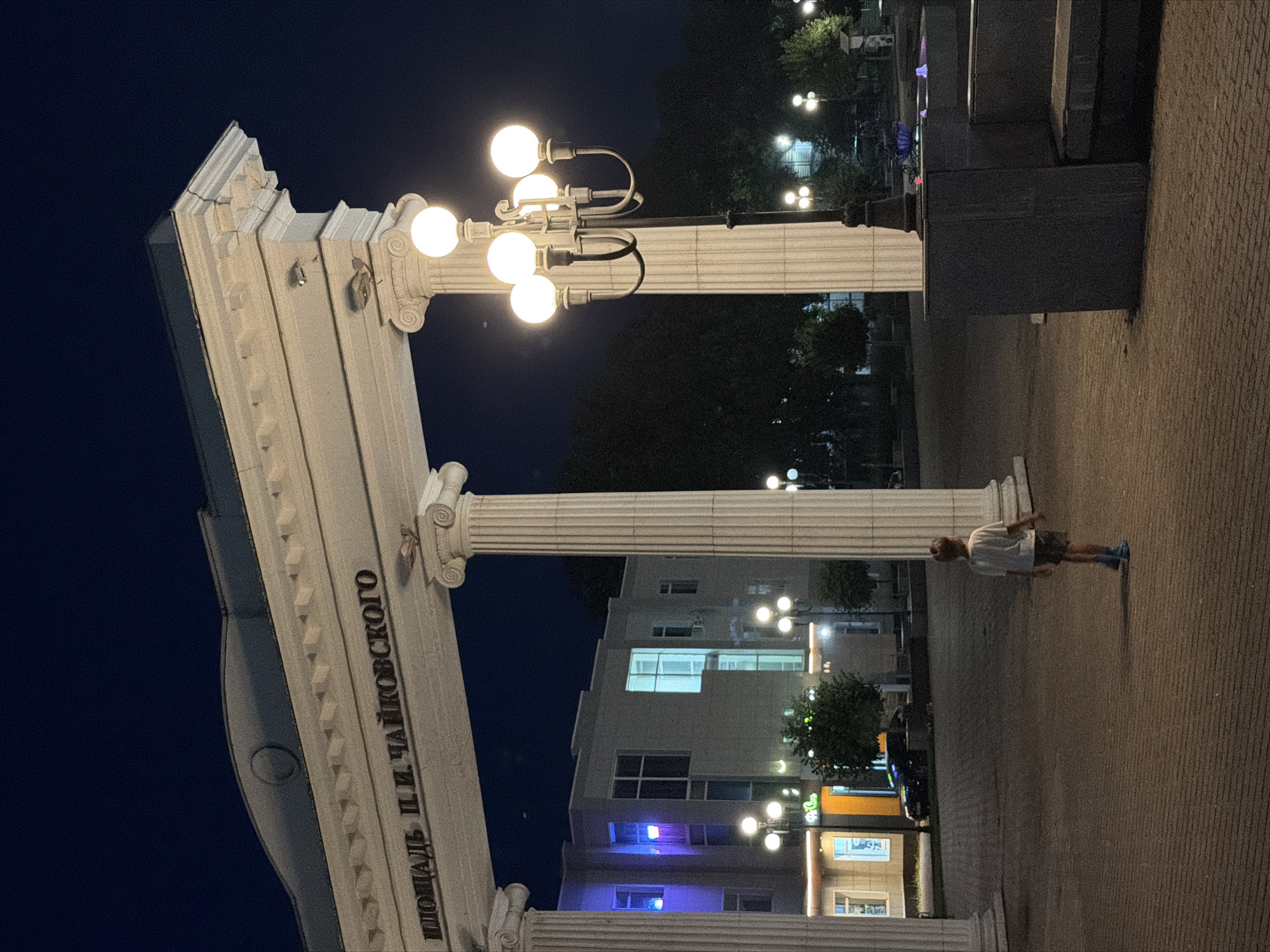 The image depicts a night scene in the city of Chaikovsky, Russia, at the square named after P.I. Tchaikovsky. A child wearing a white t-shirt and shorts stands in the foreground, facing away from the camera and looking towards a large, white, neoclassical archway with Corinthian columns. The archway has Cyrillic lettering that reads "ПЛОЩАДЬ П. И. ЧАЙКОВСКОГО". Beyond the archway, a modern building with illuminated windows is visible, along with several streetlights and trees. A multi-headed lamppost with glowing spherical lights stands to the right of the archway. The ground is paved with brick-like stones. The overall atmosphere is calm and quiet, illuminated by artificial lights against the dark night sky.