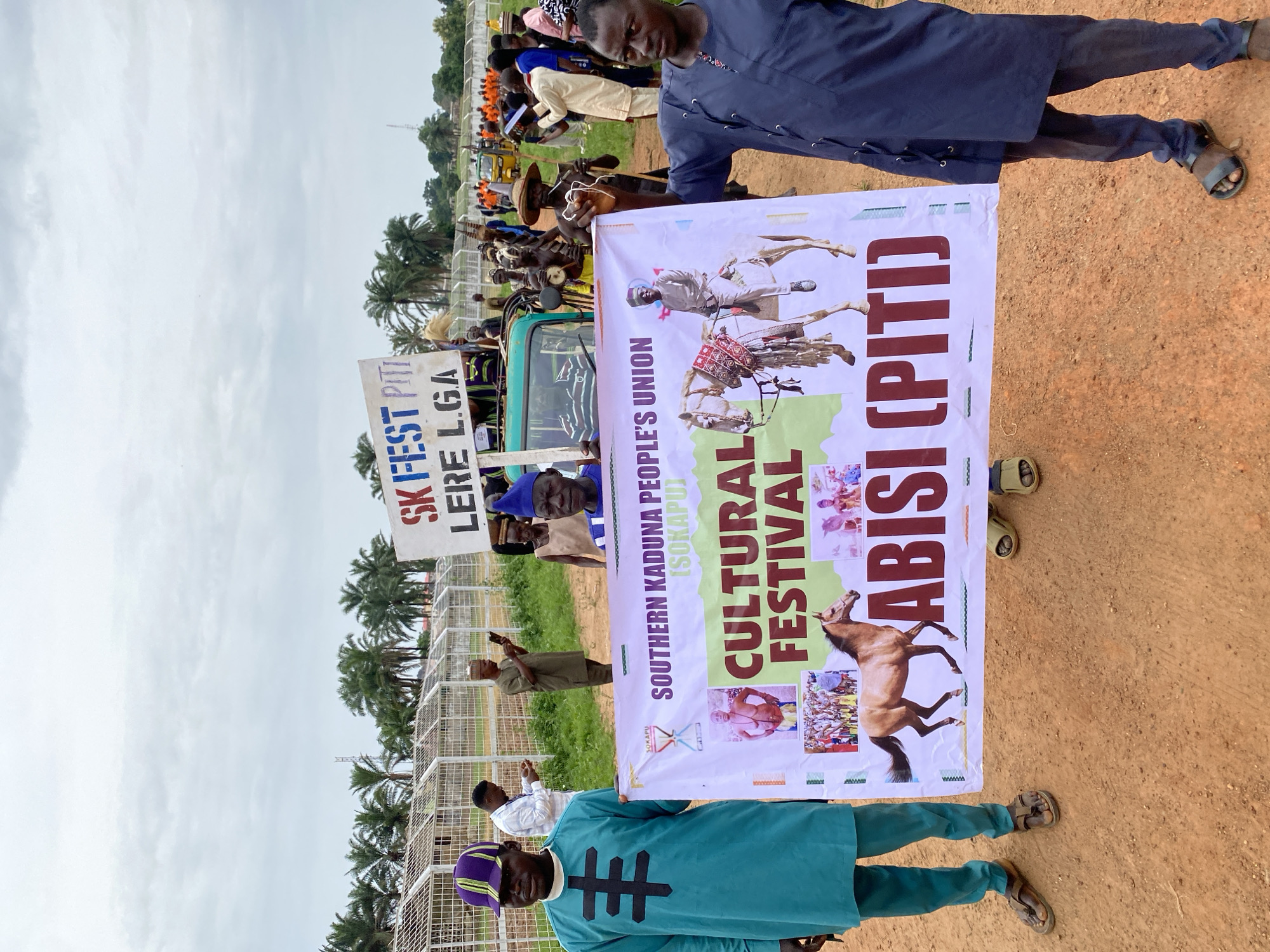 The image depicts two men holding a banner for the "Southern Kaduna People's Union (SOKAPU) Cultural Festival ABISI (PITI)". One man is in blue attire while the other wears a teal outfit with a purple cap. A second sign reads "SK Fest PITI Lere LGA". People are milling about near a green vehicle in the background on a reddish-brown, dirt covered ground. The image appears to be taken on a field with metal fencing and palm trees in the distance, suggesting it's an outdoor event. A group of musicians are seen next to the vehicle. The sky is overcast. Based on the signs, this event may be occurring in Lere Local Government Area, Nigeria.