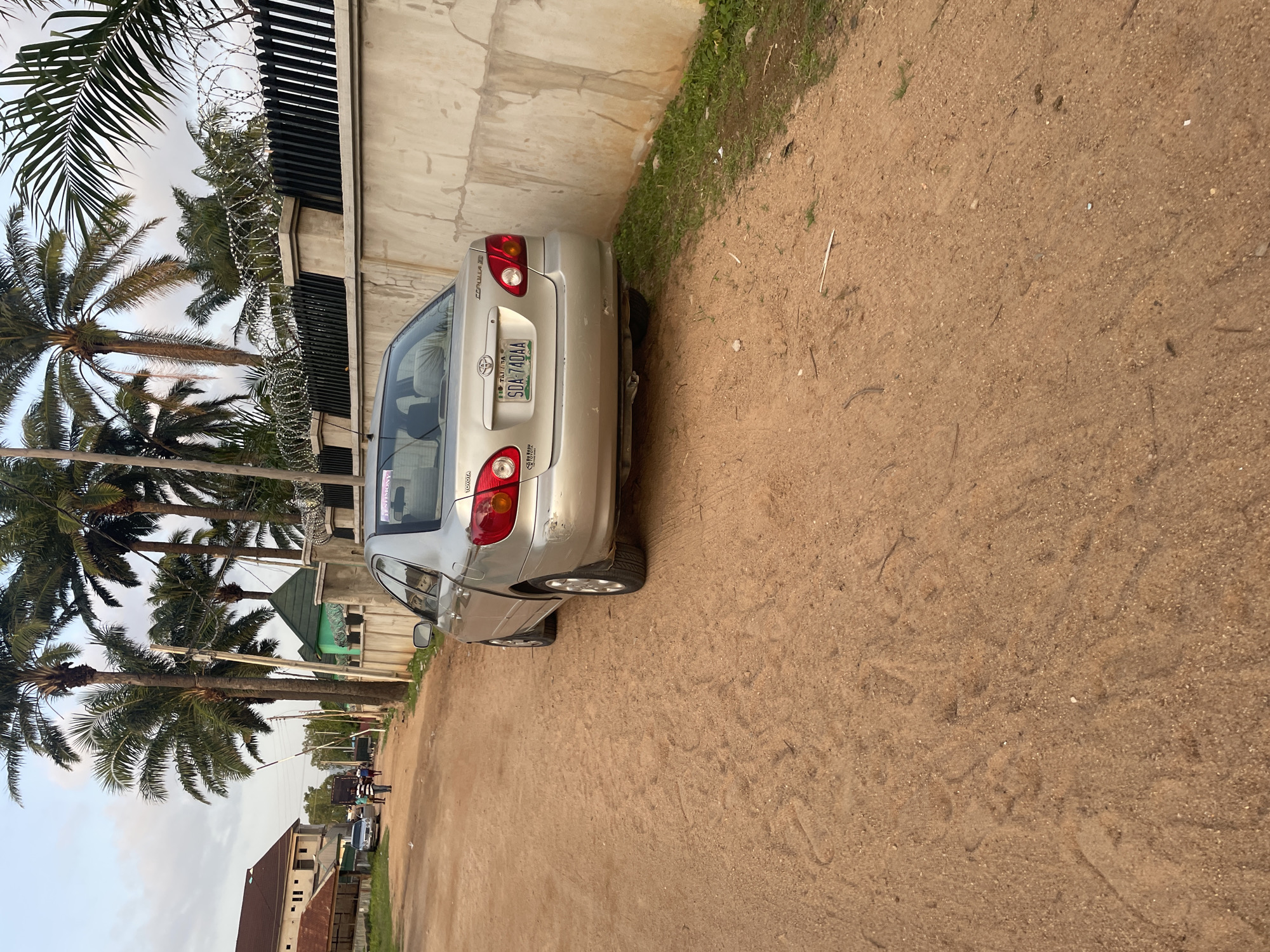 A silver Toyota Corolla is parked on a dirt road in Jalingo, Nigeria. The car is facing away from the camera, and its rear is visible. The road is unpaved and dusty, with some sparse green grass growing along the side. In the background, there is a concrete wall with barbed wire on top, a row of palm trees, and a building. A few people are walking in the distance, and another car is parked further down the road. The sky is overcast. The license plate of the Toyota Corolla is SDA 740AA.