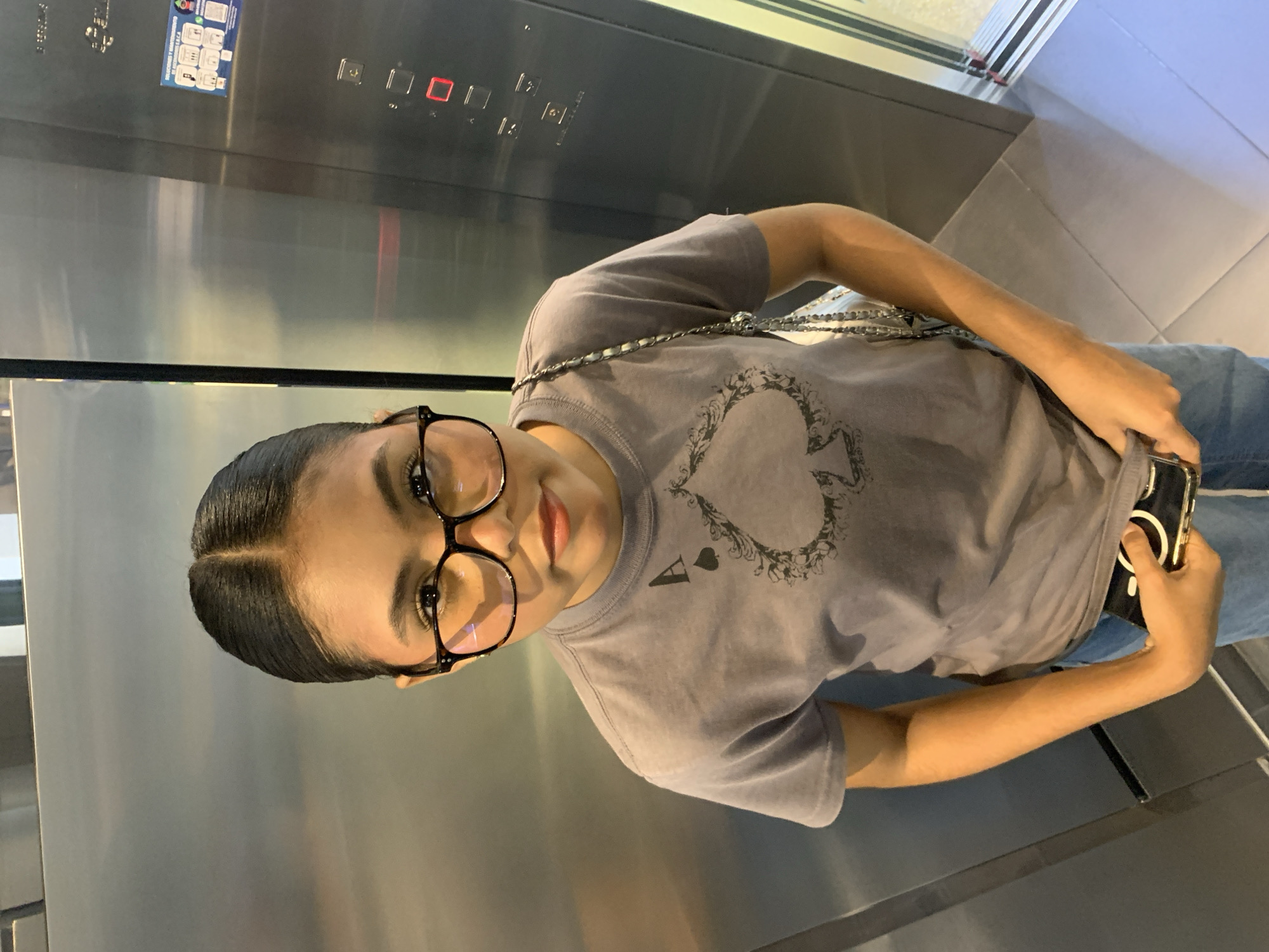 A young woman stands in an elevator. She has dark hair pulled back into a bun, and is wearing large, round glasses. Her t-shirt is grey and has a graphic of an ace of spades on it. She is holding a black smartphone in her right hand. The elevator walls are made of brushed metal, and there are control buttons on the right side of the frame. The floor is tiled. The image was taken at the Sambil shopping mall in Porlamar, Venezuela, which is a large, modern shopping center with multiple floors and a variety of stores and amenities.