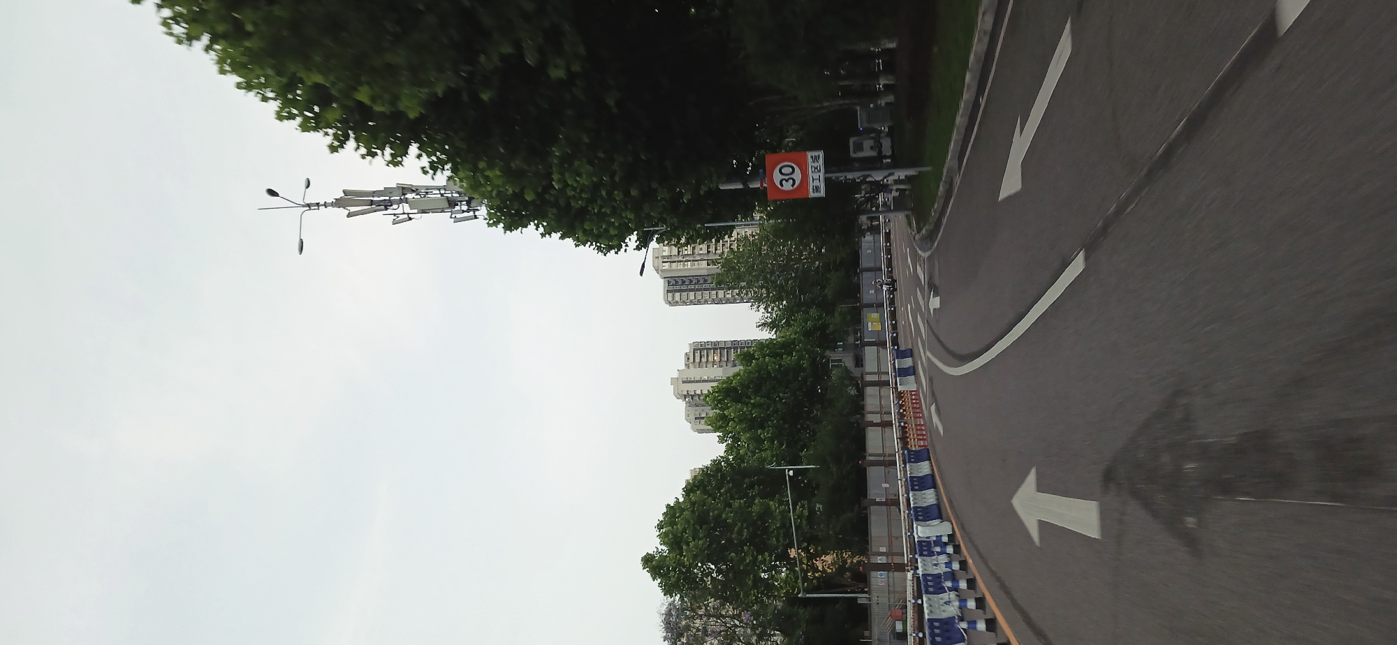 The image shows a street in Jiulongpo District, Chongqing, China. The road is paved asphalt with painted white arrows indicating direction. A speed limit sign with the number "30" is visible next to the road. There are several tall buildings in the background and trees lining the street. A street light is also visible. The scene is taken during daylight hours. There are a number of blue and white construction barriers on the side of the road.