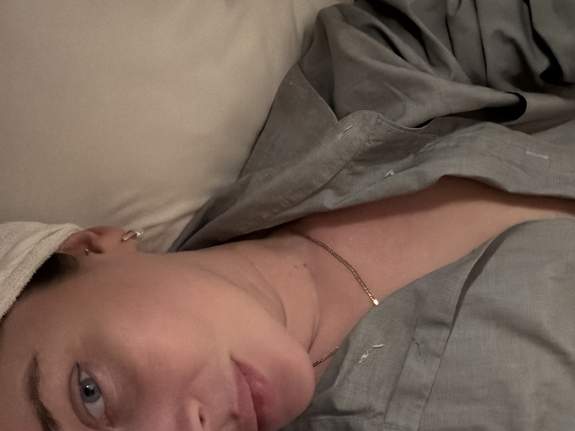 A person is lying in bed, possibly in a residential setting. The individual has fair skin, blue eyes, and a towel wrapped around their head. They are wearing a delicate gold necklace. A gray or light gray button-down shirt or pajamas covers part of their body. The bed has a light-colored pillow. There is no indication of Moscow, Russia in the image.