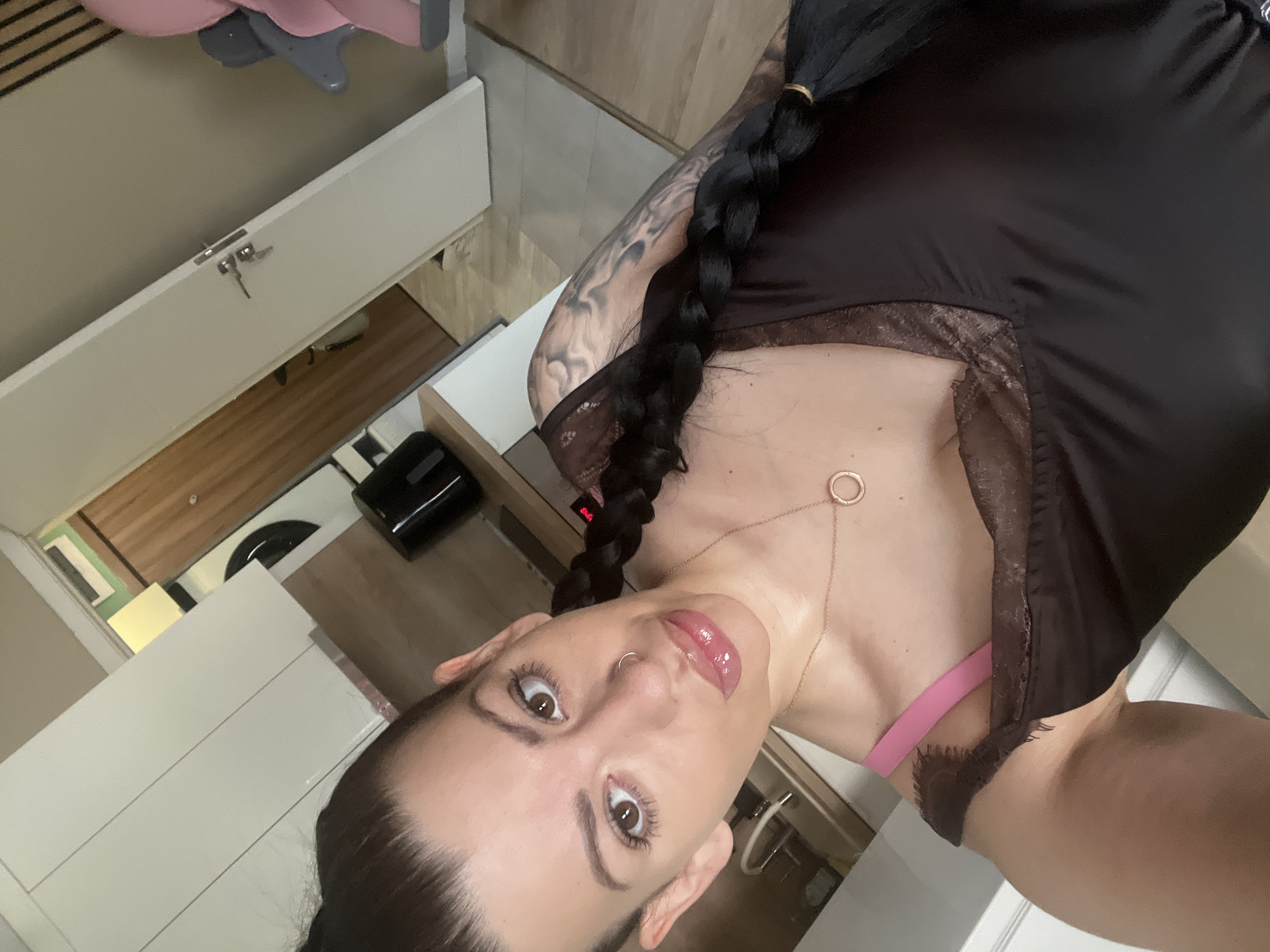 A woman is taking a selfie in what appears to be a kitchen or living area. She has dark hair styled in a braid and is wearing a dark top with lace trim and a pink bra. She is also wearing a necklace with a circular pendant and has a nose ring. The background shows kitchen cabinets, a wooden door, and a closed white door. There is also a pink and gray baby chair visible in the background. The lighting suggests an indoor setting, possibly during the day or with artificial lighting.