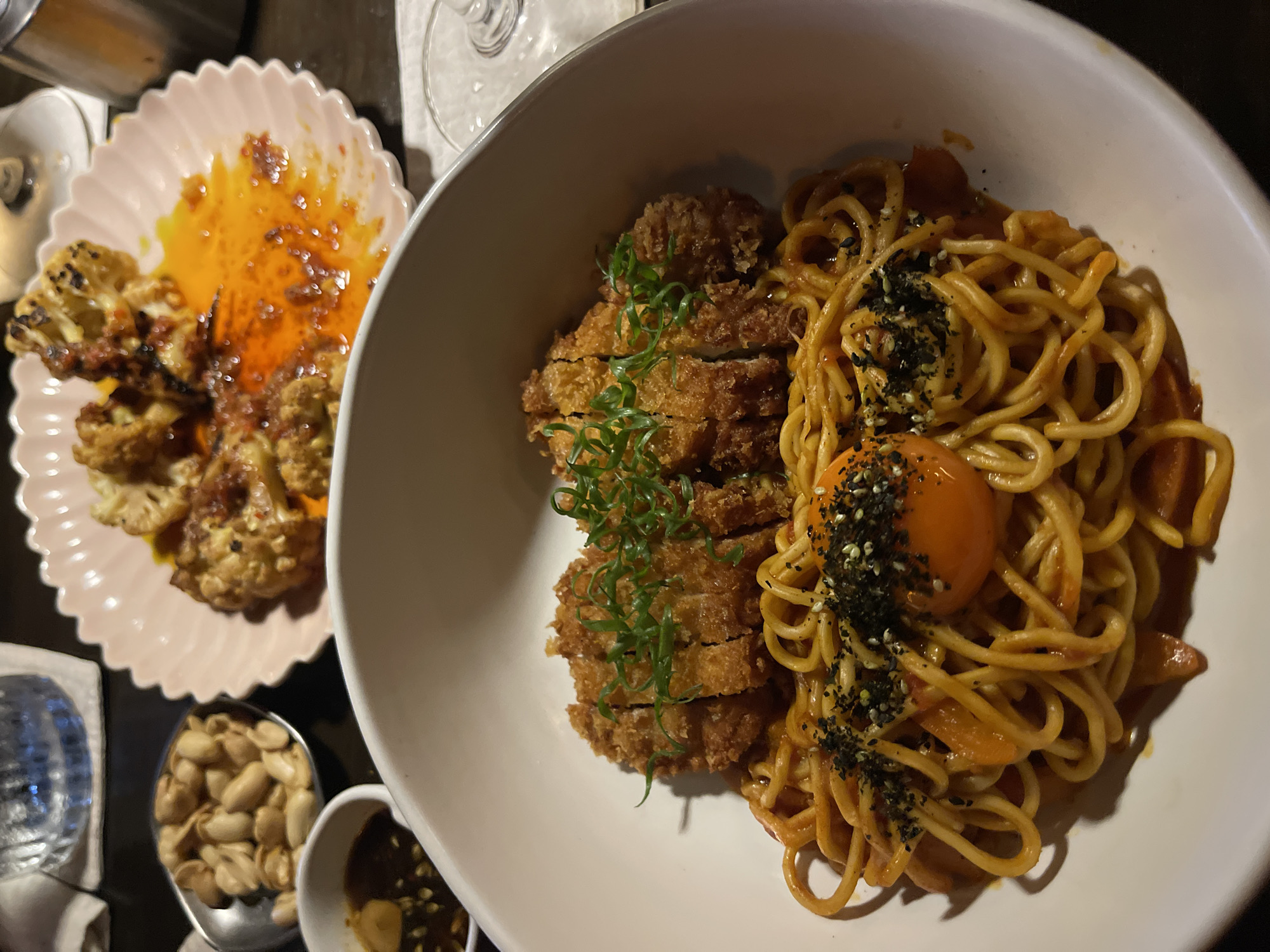 The image shows a close-up overhead view of a meal served in a restaurant in Mengwi, Indonesia. The main dish, presented in a large white bowl, consists of a portion of sliced fried chicken or pork, seasoned noodles with a reddish sauce, a raw egg yolk, and sprinkled black and white sesame seeds. To the left, on a pink fluted plate, are pieces of roasted cauliflower with a glistening orange sauce and some darker spices or herbs. In the lower left corner, a small metal scoop holds roasted peanuts, and next to it is a small white bowl containing a dark sauce, likely for dipping. The scene appears to be a dimly lit indoor setting, possibly a restaurant, with hints of glassware and other tableware visible in the background. The lighting suggests it could be evening. The focus is on the food, implying dining or preparing to dine. No people are visible, and there is no visible text in the image.