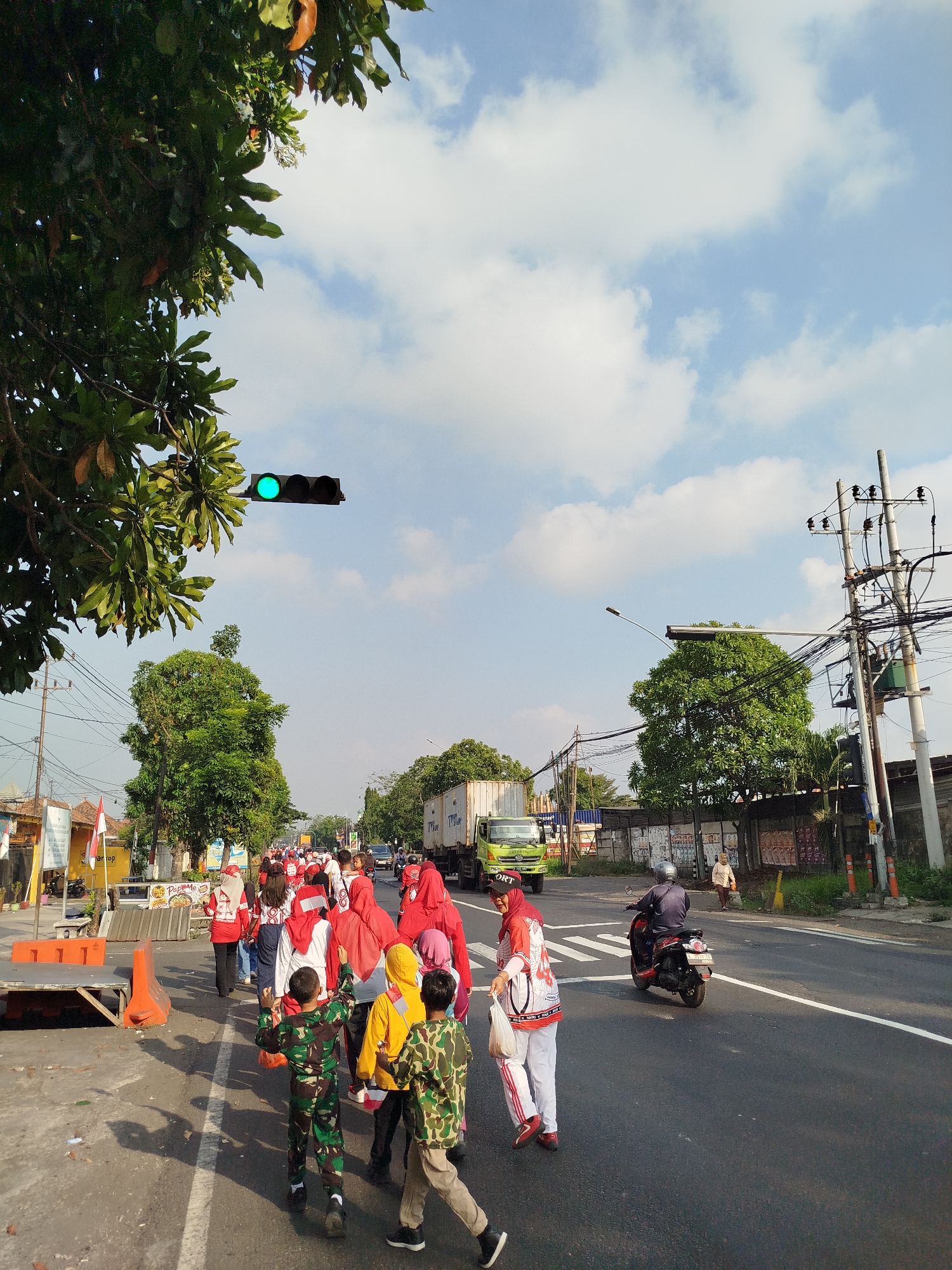 The image shows a street scene in Surabaya, Indonesia, on a bright, sunny day with scattered clouds. In the foreground, a group of people are walking along the side of the road. Some are wearing red and white outfits, suggesting a patriotic event or celebration. Among them are children dressed in camouflage military uniforms.
Further down the street, a green truck is visible, and a person on a red motorcycle is riding in the same direction. There are trees lining the road, power lines overhead, and buildings on the right side of the street. A traffic light is visible in the upper left corner, showing a green light. The road has white lane markings and a pedestrian crossing. The overall impression is of a public gathering or procession on a typical city street.
