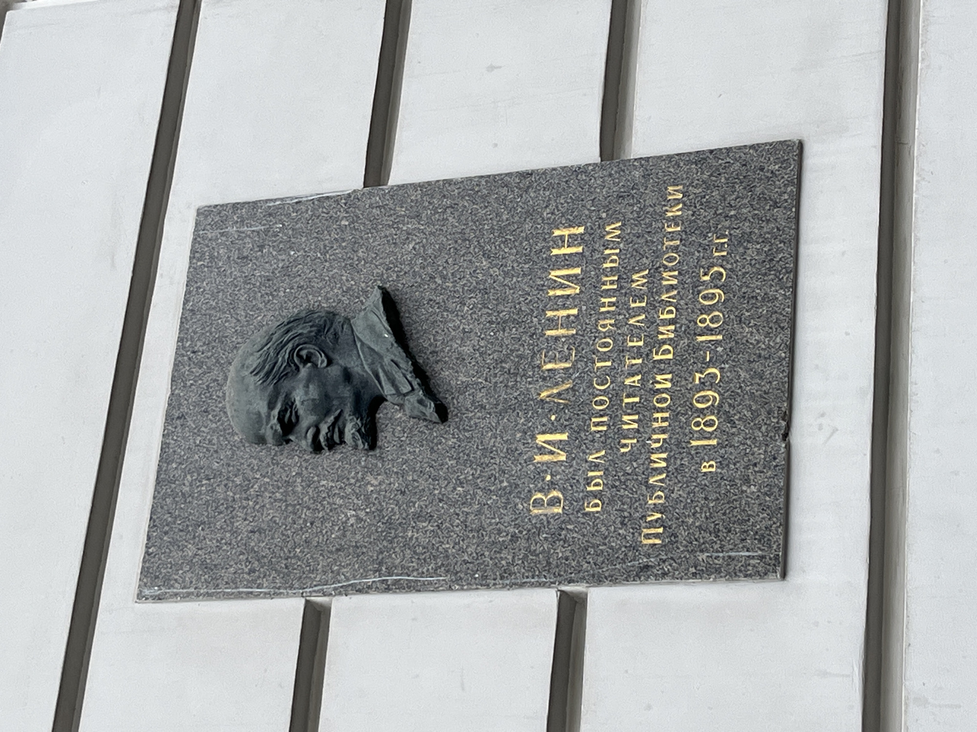 A stone plaque is mounted on a white wall with vertical grooves. The plaque is made of dark gray stone and features a bas-relief of Vladimir Lenin's head on the left. To the right of the bas-relief is text in gold lettering, written in Russian, that translates to "V. I. Lenin was a regular reader of the public library in 1893-1895." This plaque is located in Saint Petersburg, Russia.