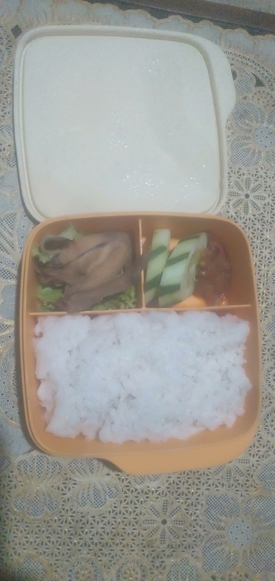 An overhead shot shows an open, rectangular, orange-rimmed bento box on a textured, cream-colored tablecloth. The bento box is divided into three compartments. The largest compartment, on the left and bottom of the image, is filled with white rice. The top left compartment contains a portion of cooked chicken served on lettuce. The top right compartment holds several slices of cucumber and a small dollop of a reddish-brown condiment. The lid of the bento box, which is off to the top of the image, is off-white and shows some water droplets. The tablecloth has a lace-like pattern with floral motifs and is a cream color with some gold thread details. The scene is indoors.
