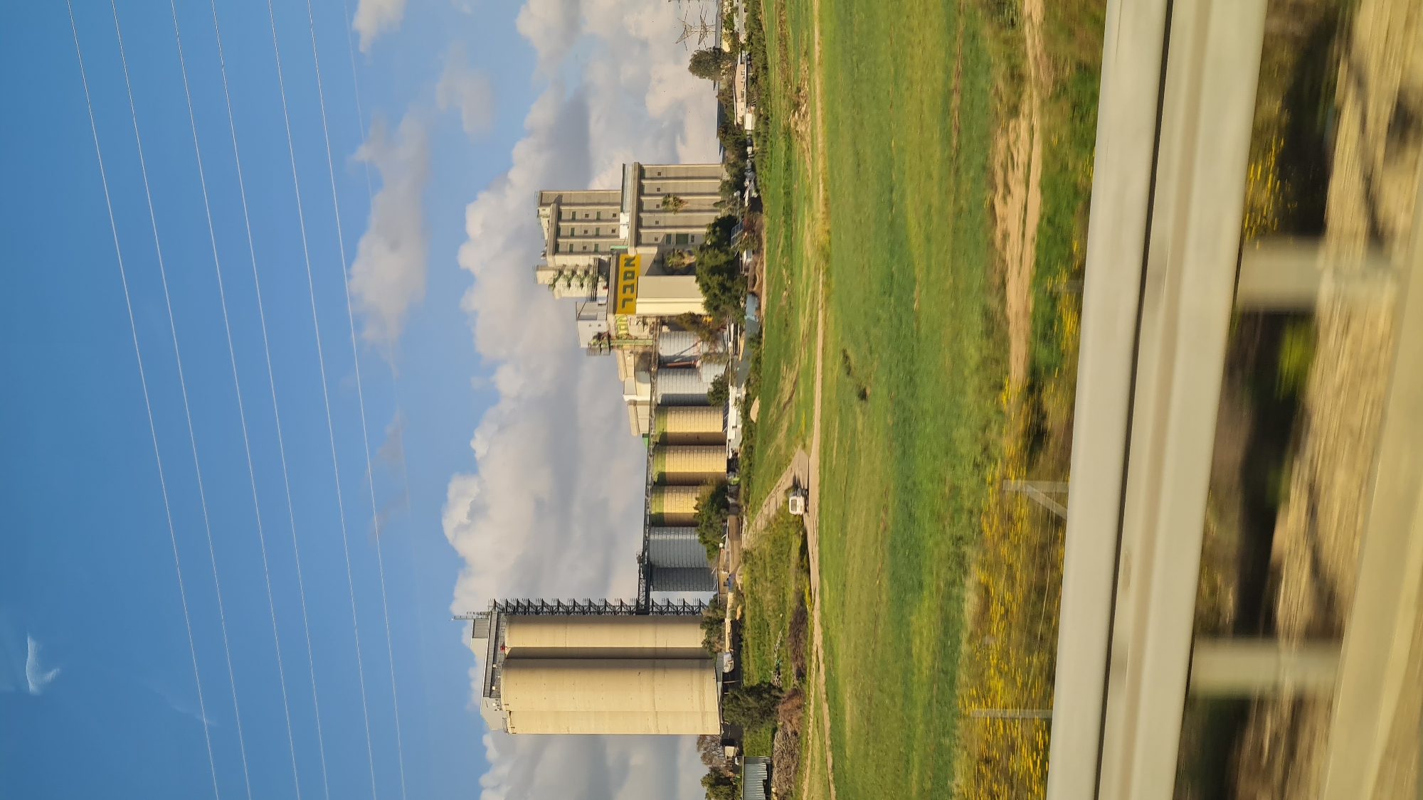 Captured from a moving vehicle, likely a train, the scene presents a landscape in Bnei Shimon Regional Council, Israel. The prominent features include an industrial complex with tall concrete silos, connected to buildings displaying the inscription "NOCL". To the right, a field with a mix of green and yellow vegetation extends, bordered by a narrow pathway. High-tension power lines stretch across a blue sky with scattered clouds on the left. The view is partially obscured by the vehicle's window frame on the right.