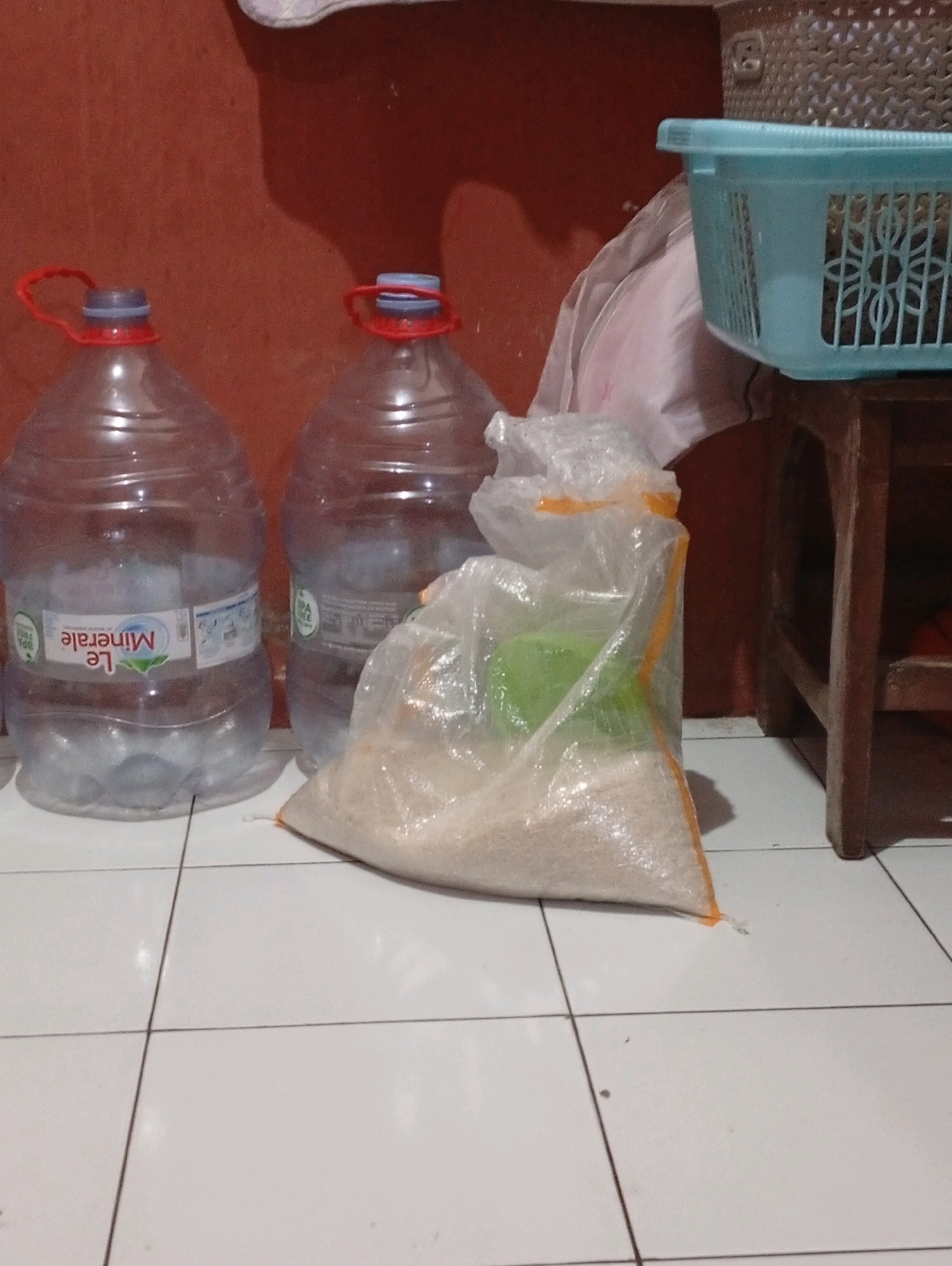 In a room with tiled flooring and a reddish-brown wall, two large plastic water bottles are placed on the floor. The bottle on the left has a red handle and a blue cap, with the label "Le Minerale" visible. The bottle on the right has a red and blue cap and a similar body. In front of these bottles, a transparent plastic bag is filled with a light-colored granular substance, possibly rice, and sealed with an orange plastic tie. A green container is visible inside the bag. To the right of the water bottles, a wooden stool supports a light blue plastic basket. The image does not provide information about the specific location within Tasikmalaya, Indonesia, or any people.