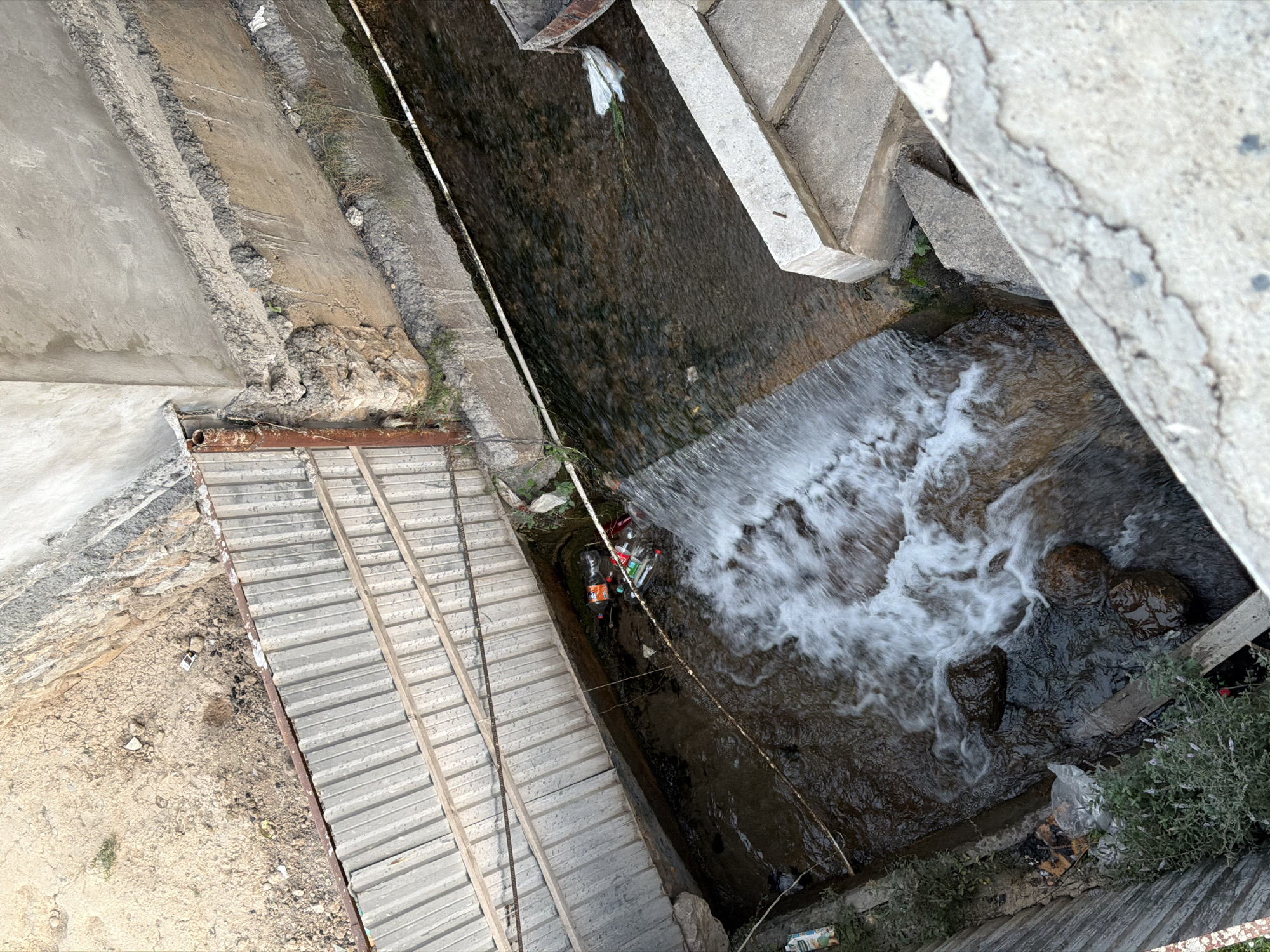 The image shows a polluted canal in the city of Nanay, Uzbekistan. The canal is filled with trash, including plastic bottles and bags. There is a small waterfall in the canal, and the water is murky and discolored. The banks of the canal are lined with concrete and corrugated metal. There are some plants growing along the banks. The scene appears to be an urban environment.
