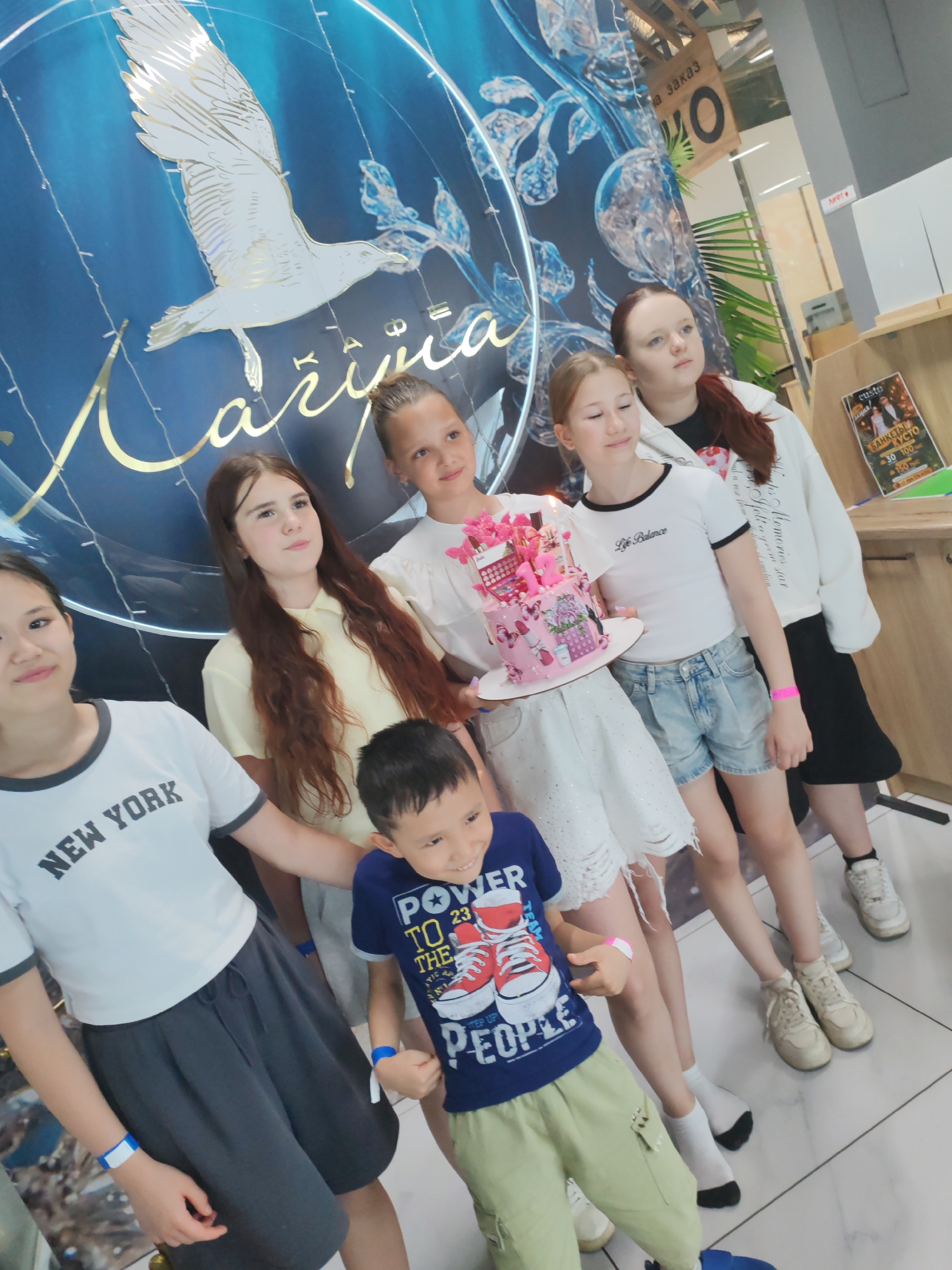 In Amursk, Russia, a group of children and young women are gathered, posing for a photo in what appears to be a brightly lit indoor venue, possibly a party hall or a shop. The group is standing in front of a large blue backdrop featuring a white seagull and stylized golden text that reads "Larisa". A vibrant pink cake, decorated with butterflies and what looks like gift boxes, is held by one of the young women, suggesting a celebration, likely a birthday.
The individuals are dressed casually. Among them are several girls and one young boy. One girl wears a white t-shirt with "NEW YORK" printed on it, paired with a dark grey skirt. Another girl in the center has long, dark brown hair and wears a yellow top. A third girl, holding the cake, is in a white top and white distressed shorts. A fourth girl, with light brown hair, is wearing a white t-shirt with "Life Balance" on it and denim shorts. The young boy in the foreground is wearing a blue t-shirt with a graphic of sneakers and text that includes "POWER TO THE PEOPLE".
Behind the group, various elements suggest a commercial or event space. There are shelves with items and posters, one of which has Cyrillic text and pricing. A sign hanging from the ceiling reads "на заказ" (for order) and "О". The overall atmosphere appears festive and joyful, with most of the subjects smiling or looking engaged, hinting at a happy occasion. The lighting is bright and even, indicative of an indoor setting.