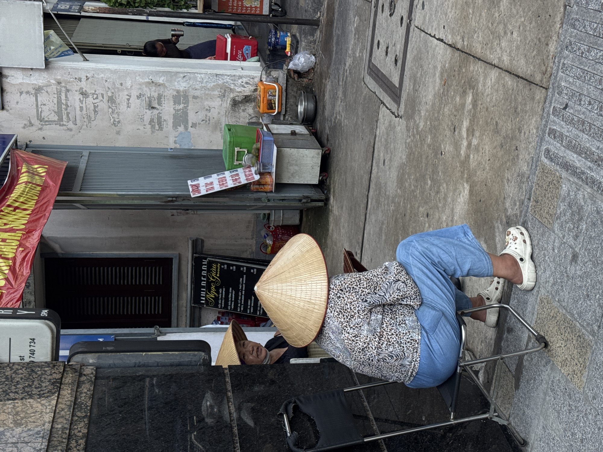 A woman wearing a conical hat sits on a metal chair, facing away from the camera. She is dressed in a leopard-print shirt and light blue pants, and white clogs. The woman is positioned on a sidewalk in front of a building. To her left, another woman wearing a conical hat is partially visible, looking towards the camera. The building features a doorway and windows with dark shutters. Various items are on display near the building entrance, including a green cooler, a blue basket with fruit, and a red cooler. A sign with Vietnamese text is visible in front of the cooler. The sidewalk is made of concrete, with a metal grate in the foreground. The scene appears to be in an outdoor urban environment.