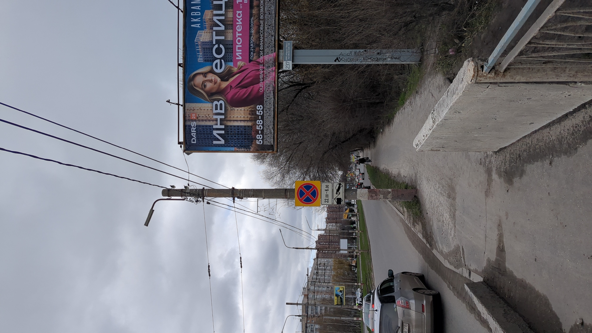 The image shows a street in Ulyanovsk, Russia. A billboard is visible on the left, advertising "DARS 25 ИНВ EСТИЦ AKBAM," which may refer to real estate. The billboard also includes details about mortgages and a phone number. Below the billboard is a "No Parking" road sign with an additional sign indicating the restriction applies from 22:00 to 6:00. A car is parked on the right side of the road. A utility pole with wires runs alongside the billboard. The street is lined with buildings, and people can be seen walking in the distance. The sky is overcast.