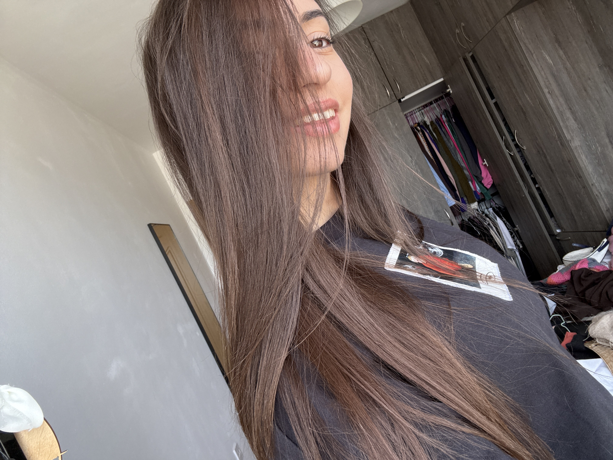 A person with long, brown hair is facing the camera, obscuring part of their face. The individual is wearing a black t-shirt with a rectangular image on it. The background features a light-colored wall, wooden cabinets, and an open closet with clothes. The location is unlikely to be Plovdiv, Bulgaria, as there is no evidence to support this claim based on the visible elements in the image.