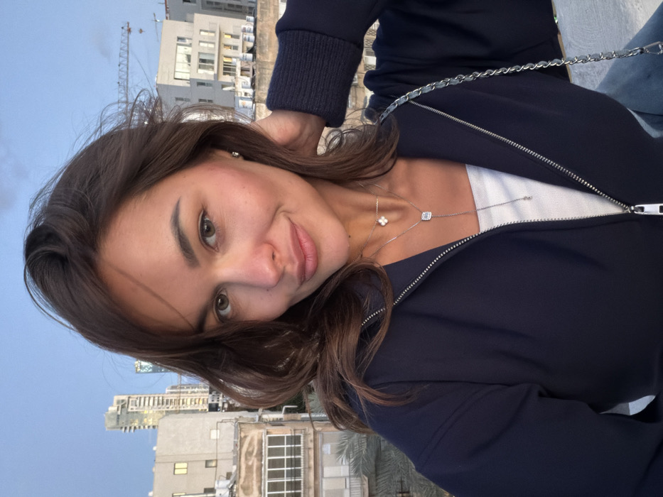 A female, appearing to be in her mid-20s to early 30s, is captured in a selfie-style shot from an elevated position outdoors. She has medium brown, wavy hair, light brown eyes, and a subtle closed-mouth smile. She wears a navy blue zippered jacket, partially unzipped to reveal a white top. Accessories include a small stud earring, a choker-style necklace with a four-leaf clover pendant, and a longer lariat necklace featuring a square clear-stoned pendant. Her right hand is raised to her hair near her right ear. A silver-toned chain from a bag is visible over her left shoulder.
The background shows an urban cityscape in Tel-Aviv, Israel. On the left, the distinctive Azrieli Sarona Tower, a tall skyscraper with vertical lighting, is prominent, indicating the view is oriented generally northeast. Below it are various older and newer buildings, some with balconies. On the right, a large construction crane is visible against the partially cloudy blue sky, alongside modern, light-colored buildings. The scene suggests late afternoon or early evening lighting.