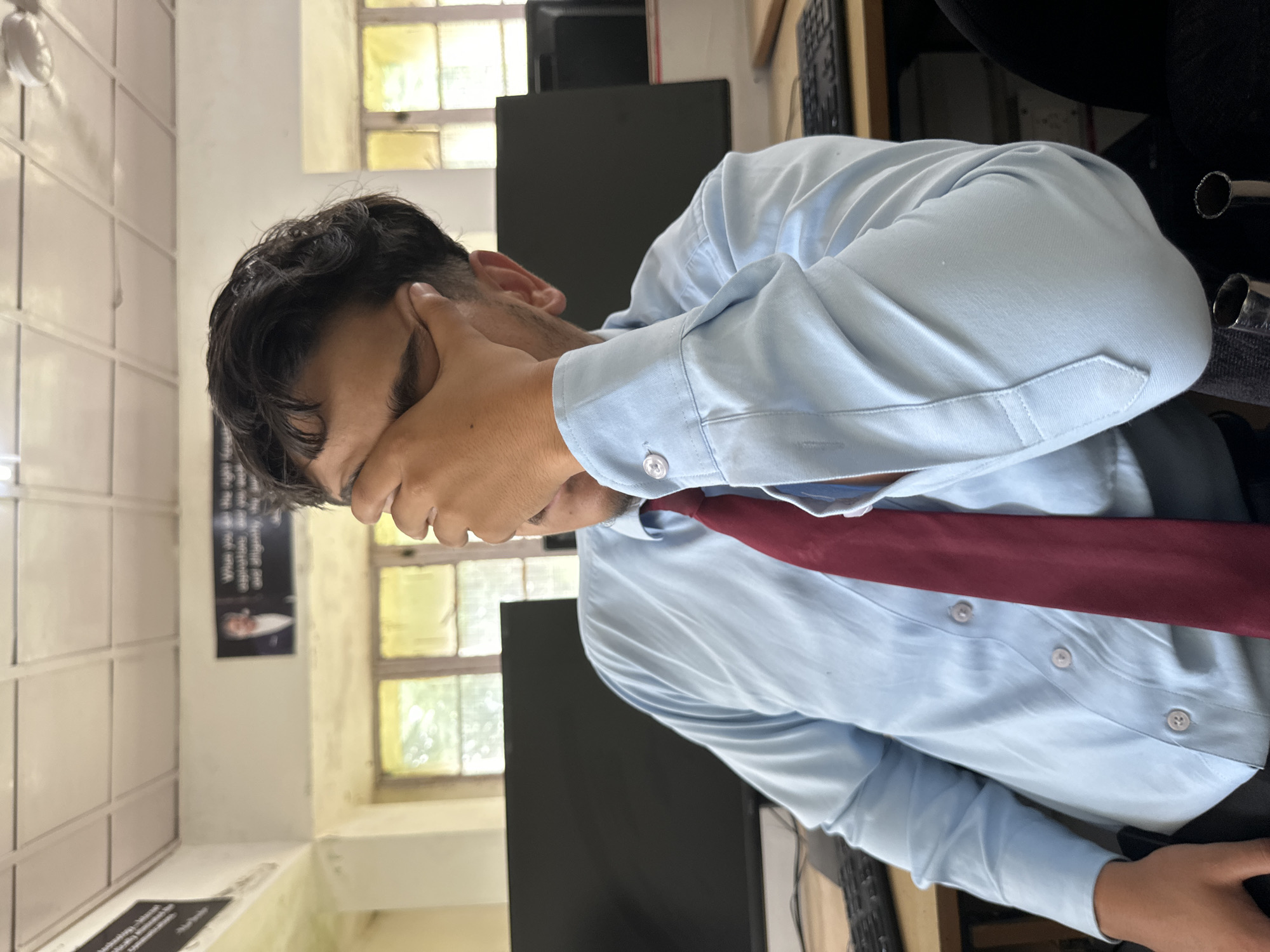 A young man with dark hair is sitting in what appears to be a classroom or office setting. He is wearing a light blue collared shirt with a maroon tie. His right hand is covering his face, obscuring his eyes and forehead, suggesting he might be tired or stressed. In the background, two black computer monitors are visible, along with a keyboard. The room has a white ceiling with fluorescent lights and a white wall. A window with yellow-tinted glass lets in natural light. On the wall above the monitors, there are two framed motivational posters. One poster features a man in a suit, and the other has text that is partially obscured. The man's left arm is resting on his lap, and his posture suggests he is slumped forward slightly. The overall scene indicates a work or study environment.