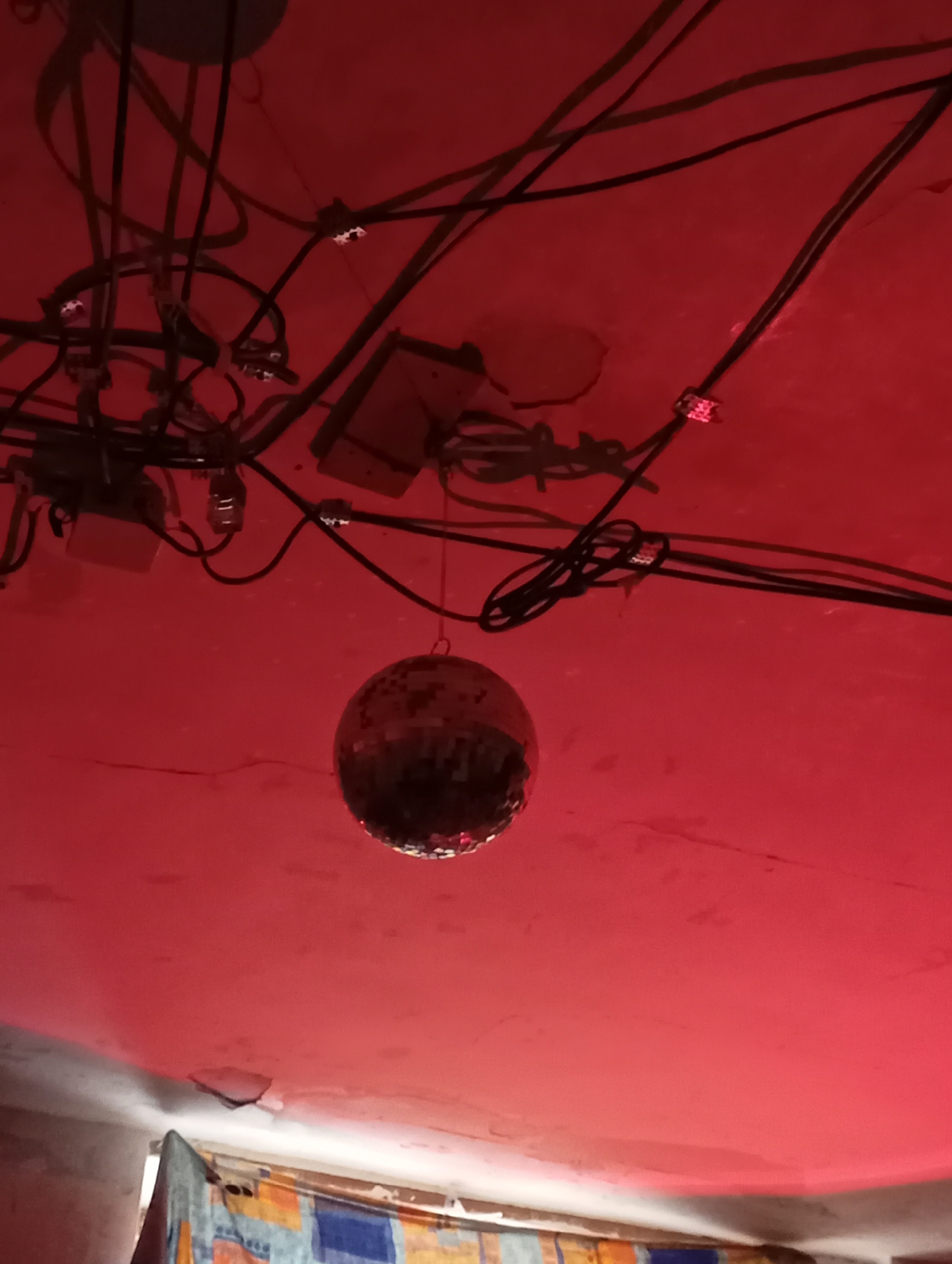 An interior scene is illuminated by a reddish light, with a disco ball suspended from the ceiling. The ceiling is cluttered with tangled black electrical wires and junction boxes. Part of a wall and window are visible in the lower left corner, with a colorful fabric or curtain. The exact location cannot be identified from the image alone. The image does not contain people. There is no evidence that the location is in the city of Krasnodar, Russia.