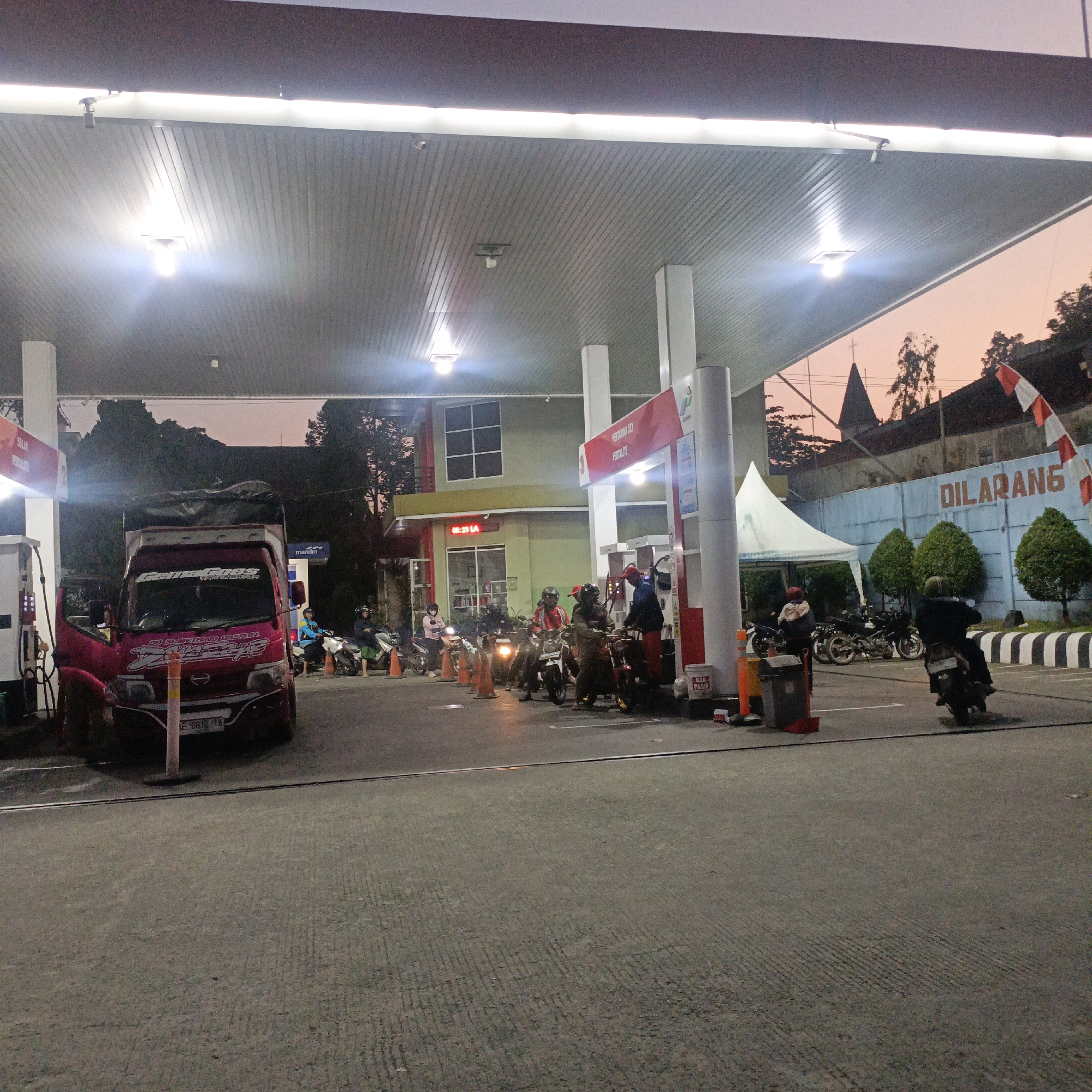 This is a photograph taken at a Pertamina gas station in Salatiga, Indonesia, at dusk. The scene depicts several people on motorcycles, some of whom are refueling. A red truck is parked on the left side of the frame, and a white tent is set up in the background. The sky is a soft orange, indicating the sun has recently set. A sign on the wall in the background reads "DILARANG," which means "forbidden" in Indonesian. There are several cones and trash cans on the ground, and the gas station is illuminated by overhead lights. The overall atmosphere is one of a busy but calm evening at a public service location.