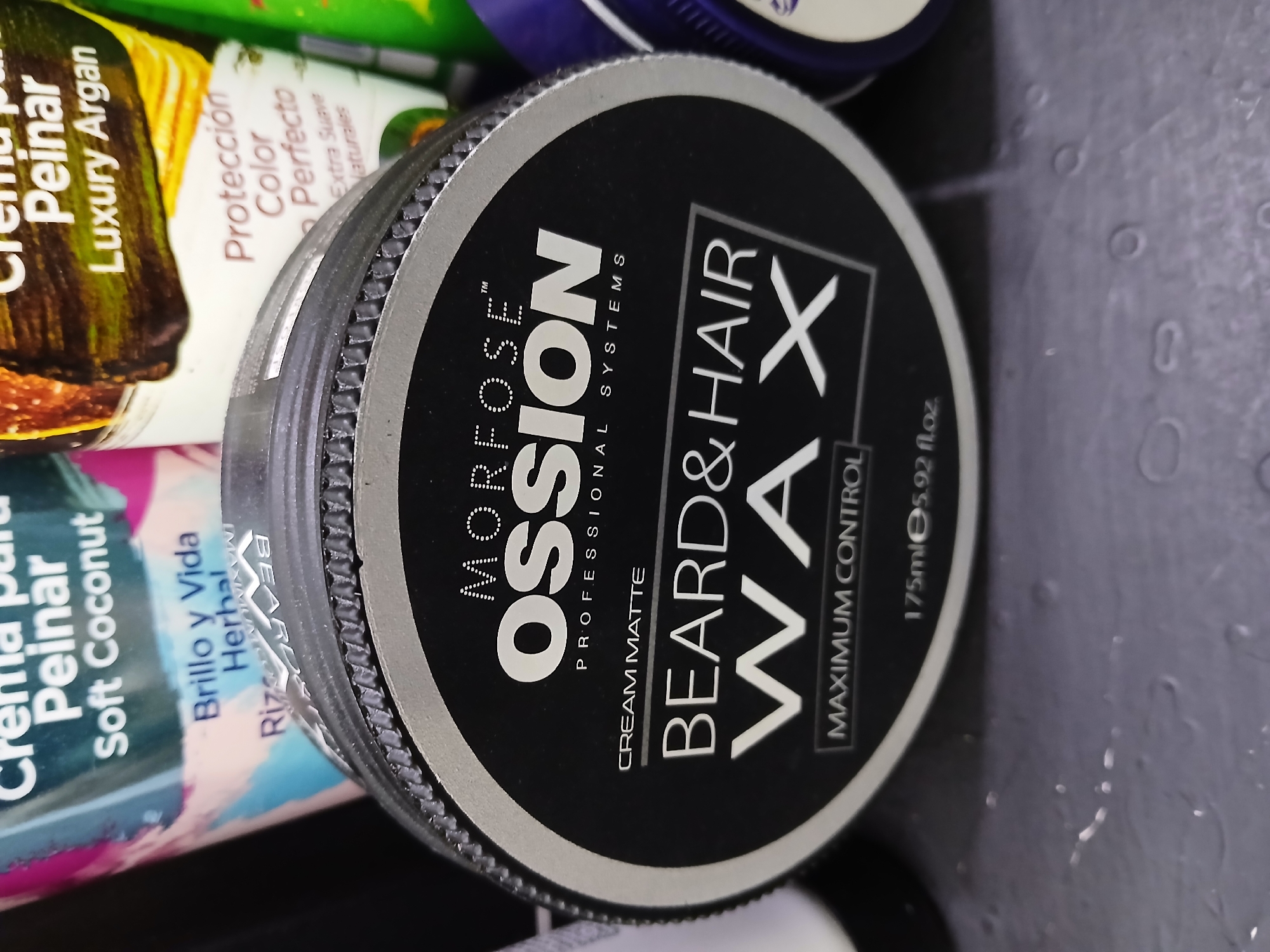 The image shows a close-up of a jar of "Morfose Ossion" beard and hair wax, with other personal care products partially visible in the background. The wax jar is black with a silver rim and contains white text that reads "Morfose Ossion Professional Systems Cream Matte Beard & Hair Wax Maximum Control 175ml 5.92 fl.oz." The background products include a hair cream labeled "Crema para Peinar Soft Coconut Brillo y Vida Herbal" and another labeled "Crema para Peinar Luxury Argan Proteccion Color Perfecto". The scene has a dark background with water droplets. The image provides no information regarding the location of Juan Griego, Venezuela.