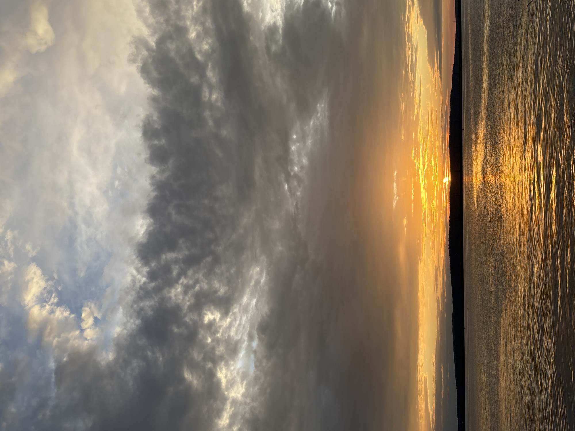 This is a vertical, medium shot of a sunset over a body of water in Chaikovsky, Russia. The sky is filled with dramatic clouds, ranging from dark gray to bright yellow and orange, with streaks of blue visible near the top. The sun is a brilliant orange ball, partially obscured by clouds, casting a golden glow on the water. The water is calm, with gentle ripples reflecting the colors of the sky. A dark, silhouetted treeline forms the horizon line, with subtle hints of distant structures. The overall atmosphere is serene and picturesque, capturing the beauty of a Russian sunset.