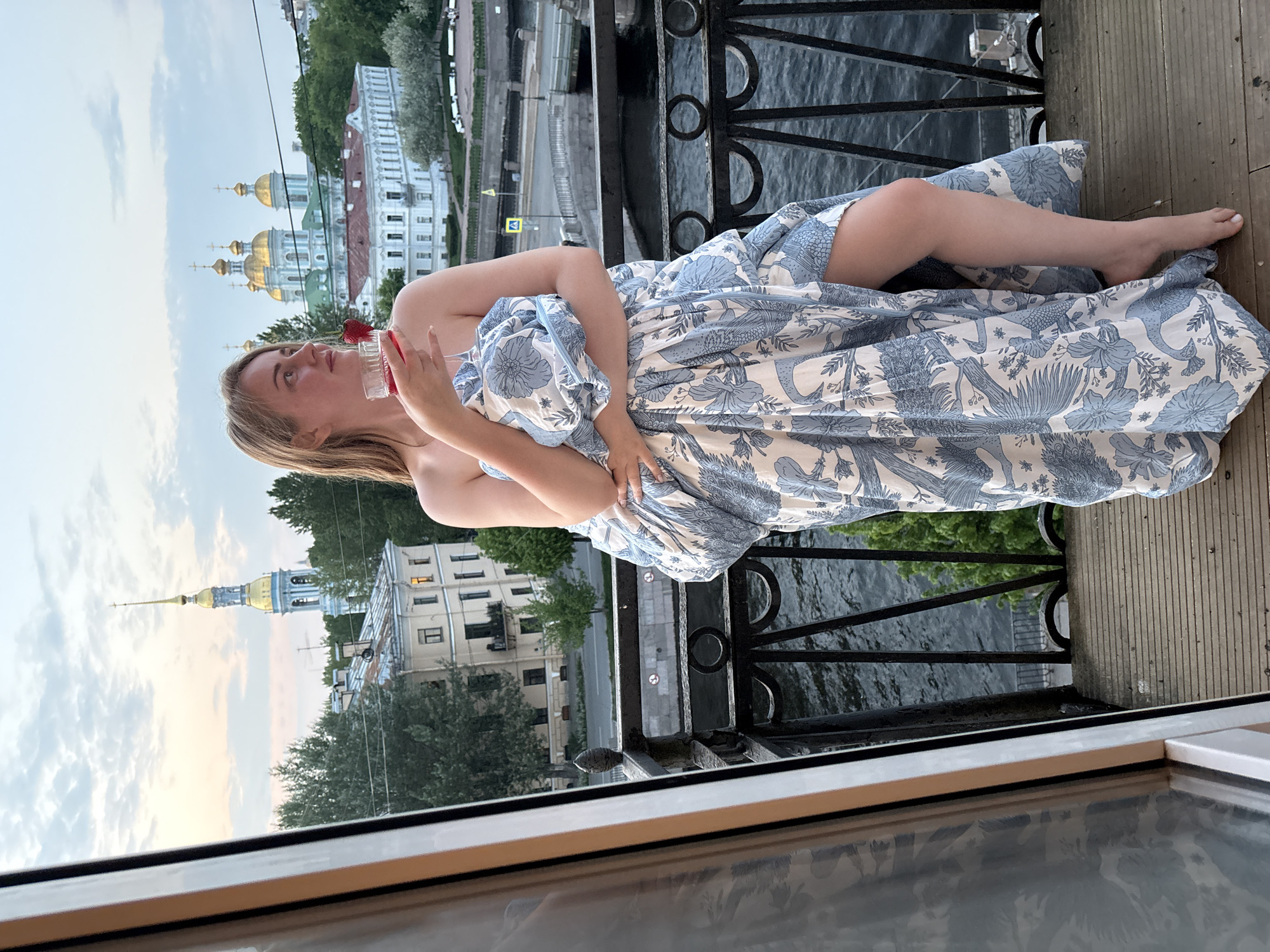 A medium shot depicts a woman reclining on a balcony or similar structure in Saint Petersburg, Russia. She holds a small dish containing strawberries. The balcony overlooks the Neva River, with views of the city's skyline including the golden domes of Saint Nicholas Naval Cathedral, and the steeple of Peter and Paul Cathedral. The woman is wrapped in a patterned blue and white fabric, with only her bare arms and one leg exposed. She is facing toward the left side of the frame, with her head slightly tilted back. The balcony has a dark-colored wrought iron railing. The lower portion of the frame is occupied by a reflective surface, possibly a window pane. The photograph is oriented in portrait, with the background skyline rotated vertically.