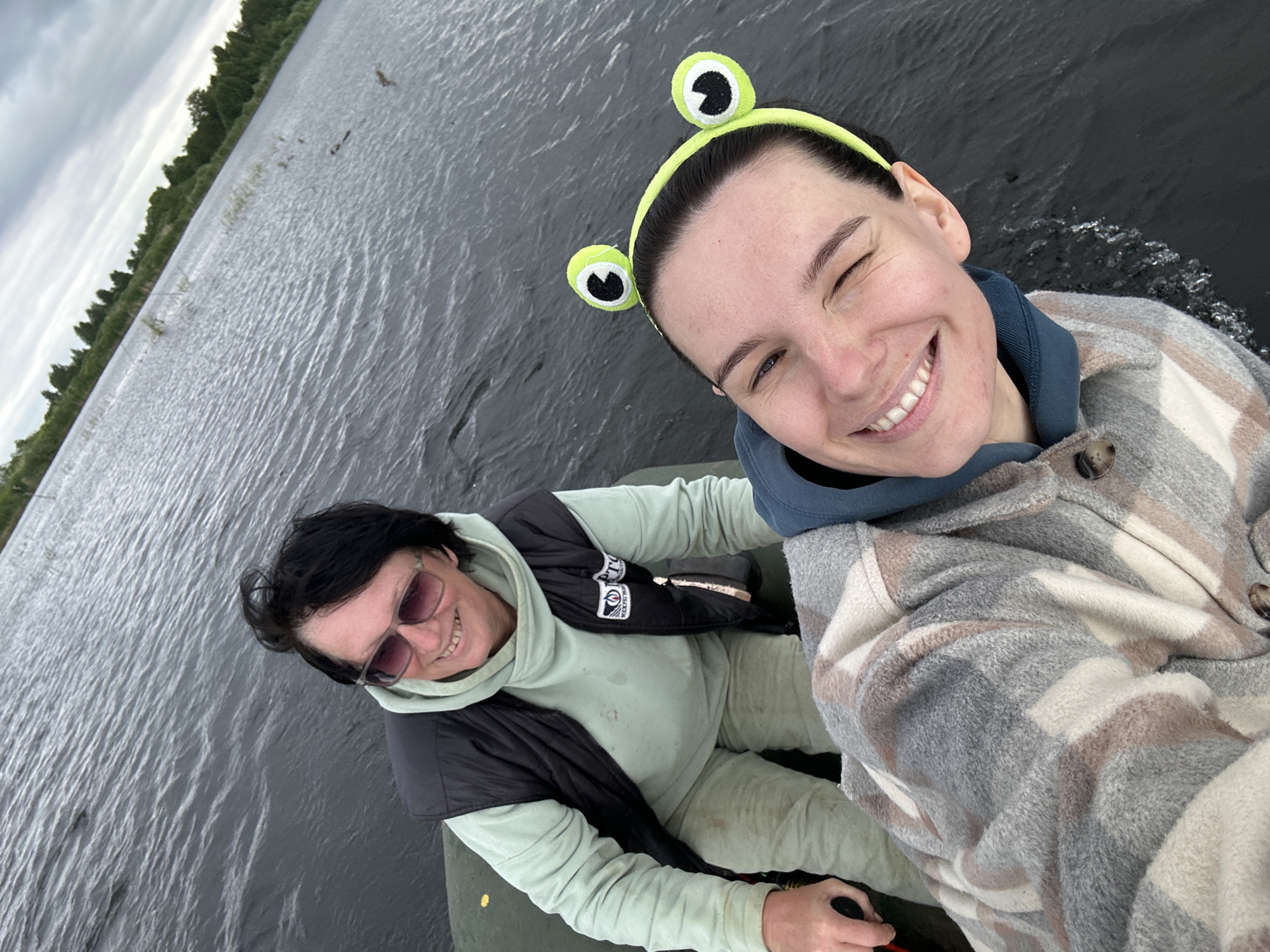 Two individuals are in a boat on a body of water, likely a lake or river, with a shoreline visible in the background. The person in the foreground is a woman with dark hair wearing a grey and white plaid jacket and a blue hooded sweatshirt. She is smiling and has a frog-themed headband on. The person behind her is an older woman with dark hair, wearing sunglasses, a black vest over a light green hooded sweatshirt, and tan pants. She is also smiling. The water appears choppy, and the sky is overcast. The location is identified as Dubrova, Russia.