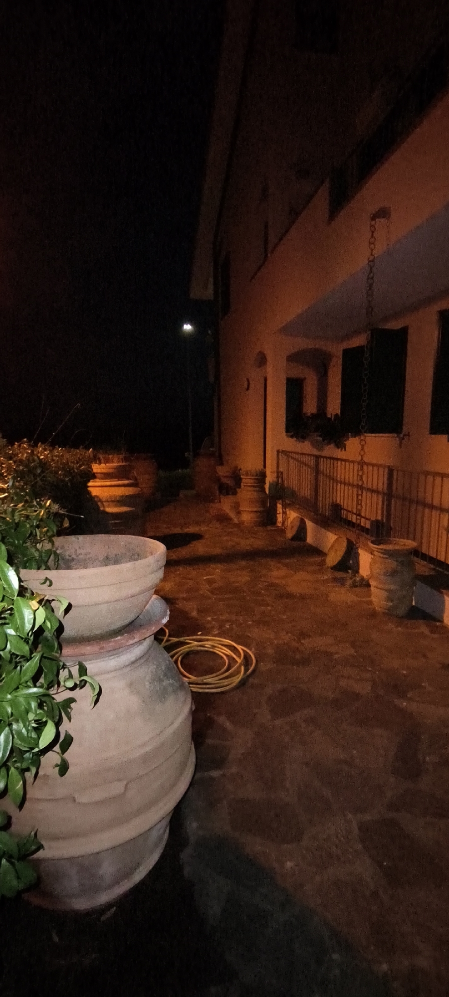 This is a nighttime, full-shot, outdoor photograph taken in Campagnatico, Italy. The scene depicts a paved patio or walkway in front of a building, illuminated by artificial light. In the foreground, to the left, are two large, terracotta pots, one stacked on top of the other. The top pot is empty and appears to be a planter. Lush green foliage spills over the rim of the top pot. To the right of the pots, a yellow garden hose is coiled on the ground.

The building, situated on the right side of the frame, is a multi-story structure with an orange-tinted facade. It features a balcony or porch area with a railing. Several windows with dark shutters are visible on the building's upper level. A chain hangs from the facade above the balcony.

Further into the background, a street light illuminates a dark, open area, possibly a garden or unlit yard, to the left of the building. The ground surface is paved with irregular stones or tiles, and shadows are cast by the building and objects. Several smaller, cylindrical objects that resemble barrels or planters are placed along the patio. The overall impression is one of an outdoor space at night, with some decorative elements and functional items visible.