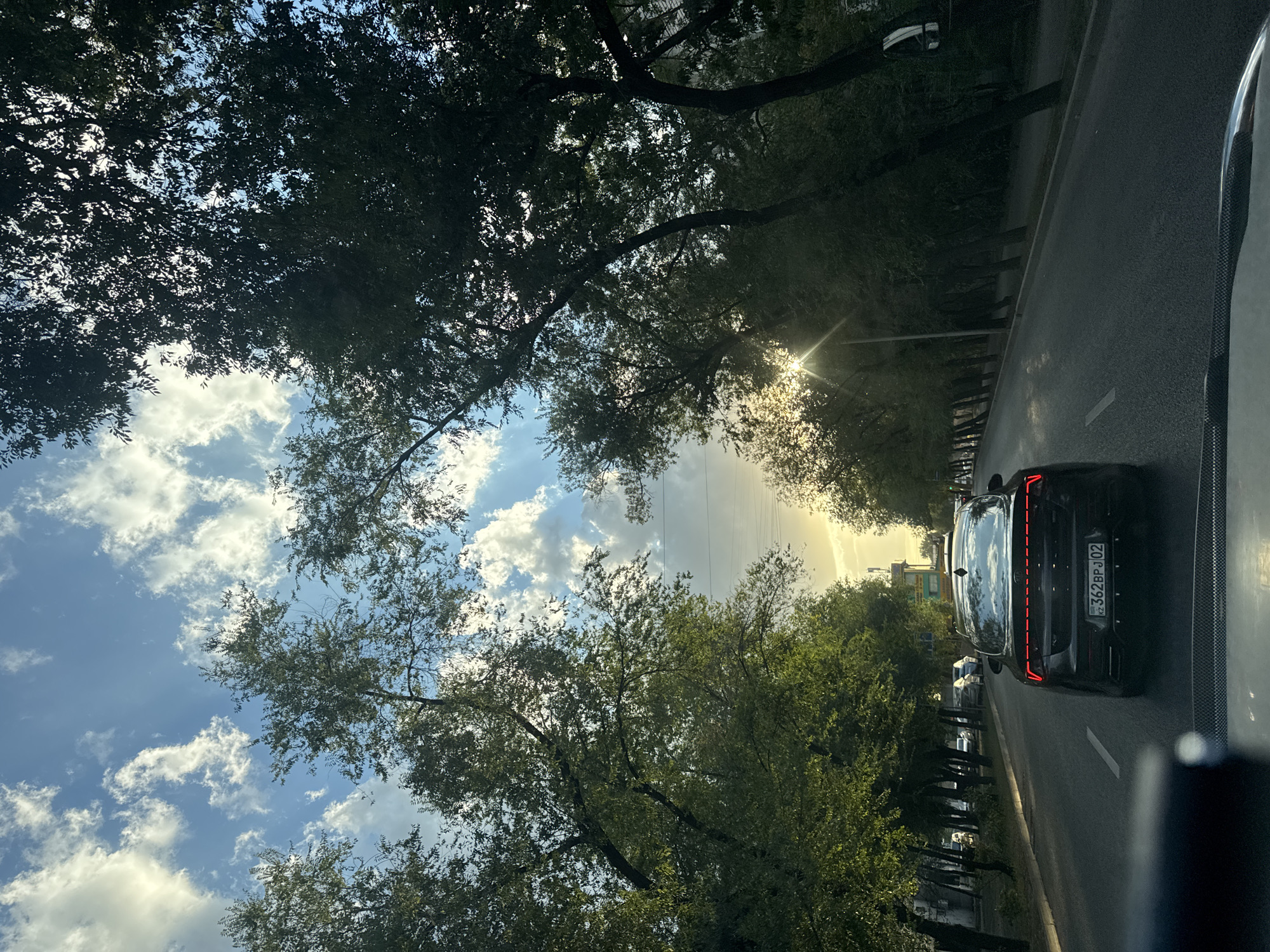 A black Kia K5 sedan is driving down a tree-lined street in Almaty, Kazakhstan. The car has a license plate reading "KZ 362 BPJ 02". The sun is shining through the trees, creating a lens flare effect. The sky is blue with scattered white clouds. The street appears to be an urban area with buildings visible in the background. The image is taken from the perspective of another vehicle.