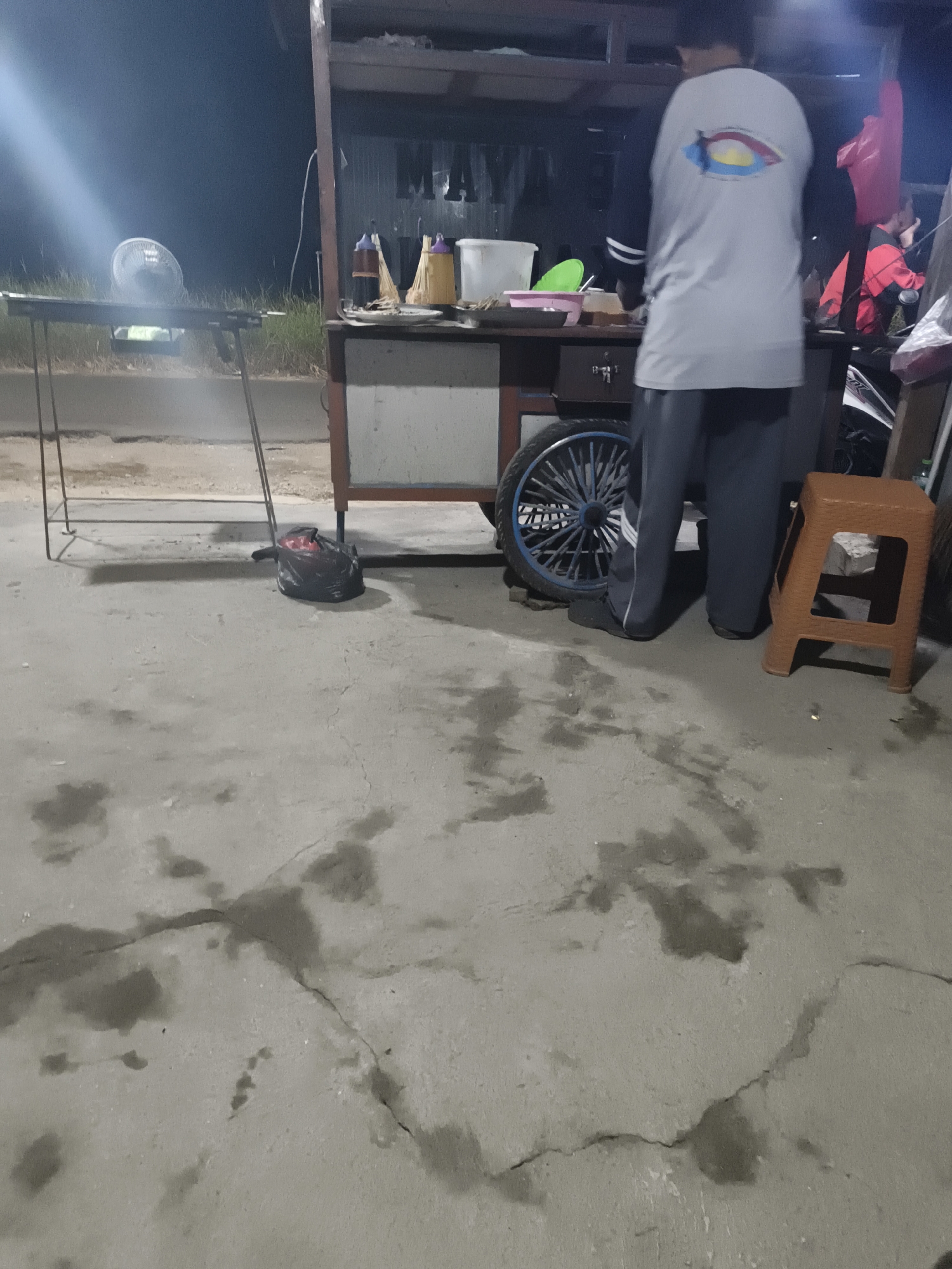 In Sidorejo, Indonesia, at night, a person stands behind a food cart. The cart has a wheel and shelves displaying food items and condiments. A fan is positioned on a metal rack to the left of the cart, illuminated by a bright light. A black trash bag lies on the concrete ground near the cart. The ground itself is uneven, with visible cracks and dark, wet patches. In the background, other people and a motorbike are partially visible, suggesting a street-side stall or market setting.