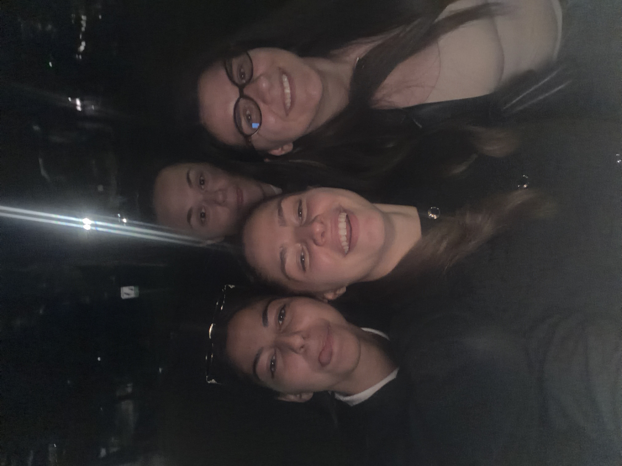 This close-up group selfie features four women in a dimly lit environment, likely at night in Etropole, Bulgaria. The image is oriented sideways, with the subjects appearing to look up at the camera.
In the foreground, one woman is visible, playfully sticking her tongue out. Above her, another woman smiles broadly with teeth showing, radiating cheerfulness. To the left and slightly behind her, a third woman with dark hair and glasses smiles widely, also showing her teeth. Her glasses reflect some light. The face of the fourth woman is partially visible on the far left, looking towards the camera.
The background is dark and indistinct, suggesting an indoor setting or an outdoor night scene with minimal ambient light. Several bright, horizontal streaks of light cut across the left side of the frame, possibly reflections from windows or distant light sources, adding to the low-light atmosphere. The overall mood is jovial and relaxed, indicative of a friendly gathering. No discernible text is visible in the image.