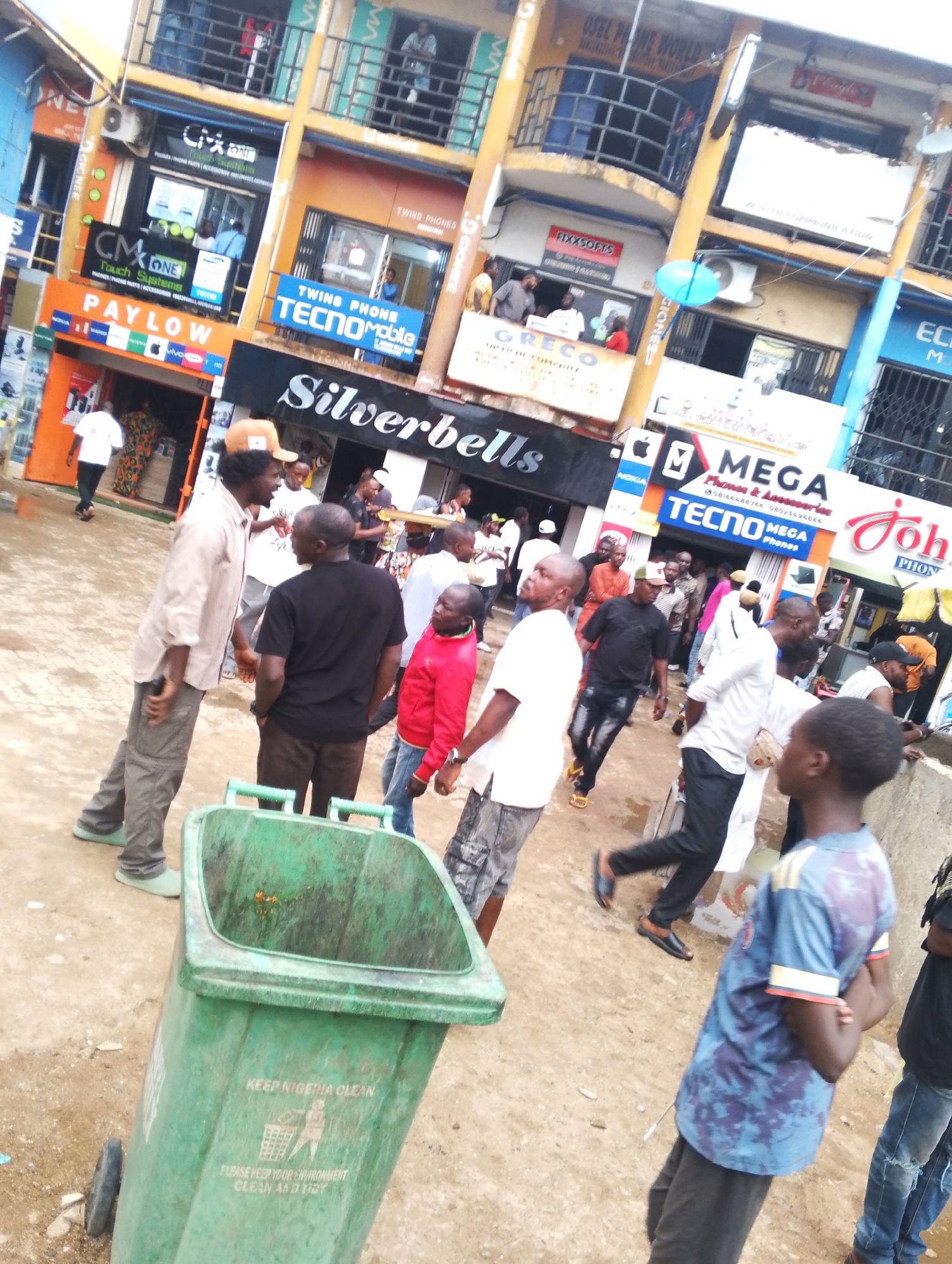 The image shows a busy street scene in Jos, Nigeria. Several people are walking around, some interacting with each other. Shops line the street, advertising mobile phones and accessories. Visible shop names include "Silverbells," "MEGA Phones," "Tecno Mobile," and "CMX One Touch Systems." A green, wheeled trash container with "KEEP NIGERIA CLEAN" printed on it is in the foreground. The ground appears wet. The buildings are multi-story with various architectural details.