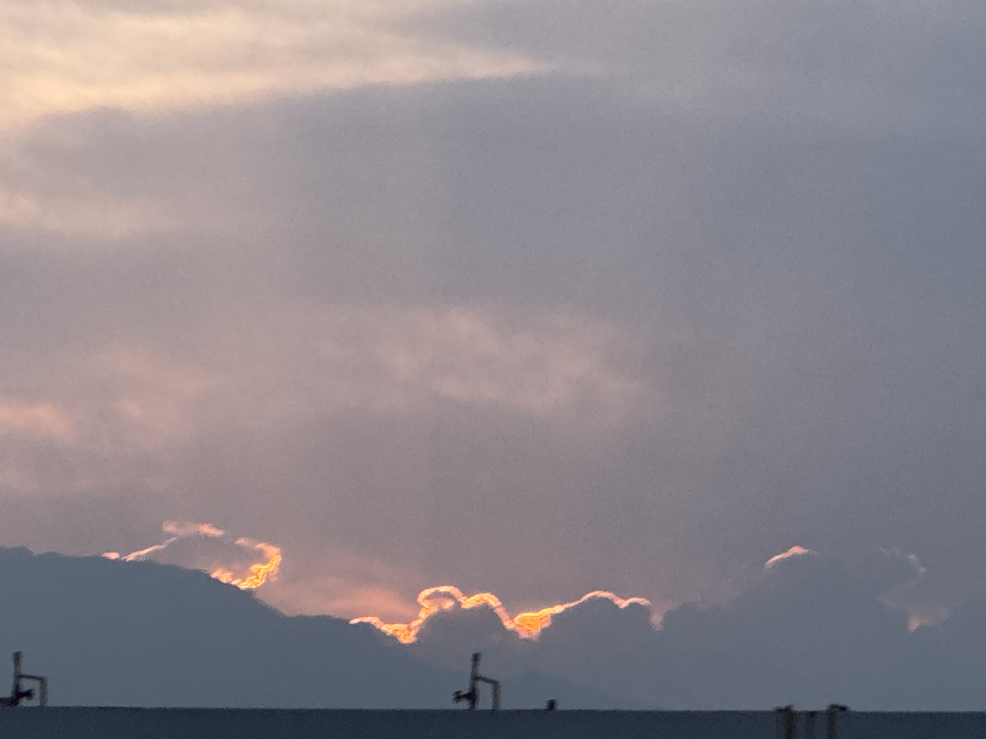 The image shows a sky with clouds illuminated by a setting sun. The clouds are a mix of dark gray and light pink. The sunlight highlights the edges of the clouds. In the foreground, there is a dark gray horizontal line, likely the top of a building or structure. There are some antennas or poles on top of the building. The scene is likely from the city of San Francisco Ocotlán, Mexico.