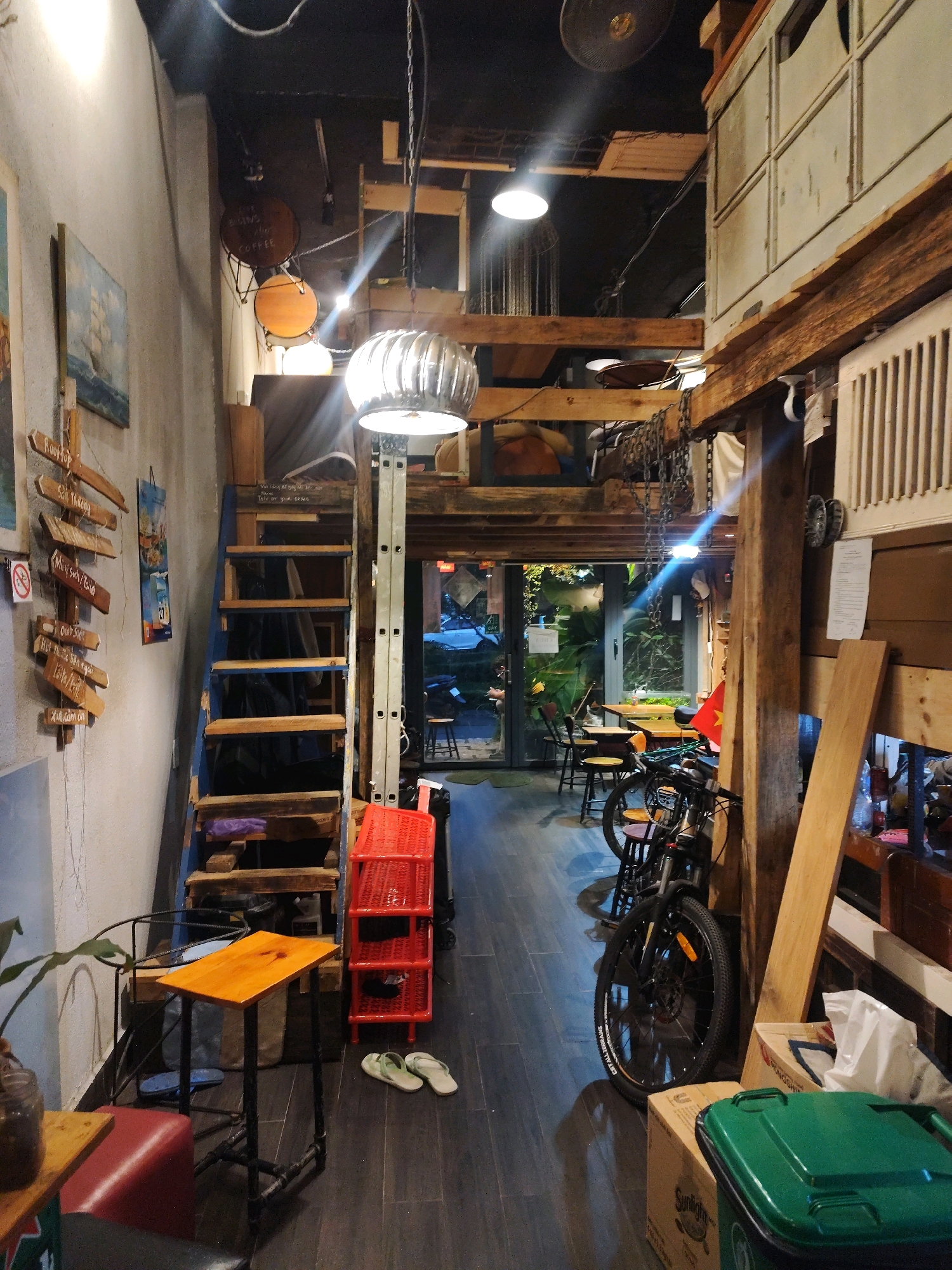 The image depicts the interior of a cafe in Da Lat, Vietnam, characterized by a rustic, industrial, and bohemian aesthetic. The space features exposed wooden beams, a mezzanine level accessible by a steep wooden staircase, and an abundance of eclectic decor. Hanging from the ceiling are industrial-style lamps, including a prominent metallic fan-shaped light fixture. The walls are adorned with wooden signs, paintings, and various hanging objects.
In the foreground, to the left, a small orange wooden table with a metal pipe base is positioned near a red cushioned stool. A plant in a glass jar sits on another low wooden table to the left. Adjacent to the stairs is a bright red plastic shelving unit. On the floor, a pair of light green flip-flops are visible.
The staircase leads up to the mezzanine, where a dark draped material is visible. A bicycle is parked vertically against a wooden beam on the right side of the image, with its front wheel facing the viewer. Further back, through an open doorway, tables and chairs are set up in an outdoor seating area, with lush greenery visible beyond. The floor is covered with dark wooden planks. The overall impression is of a cluttered yet charming space, likely a popular spot for young people due to its unique design.