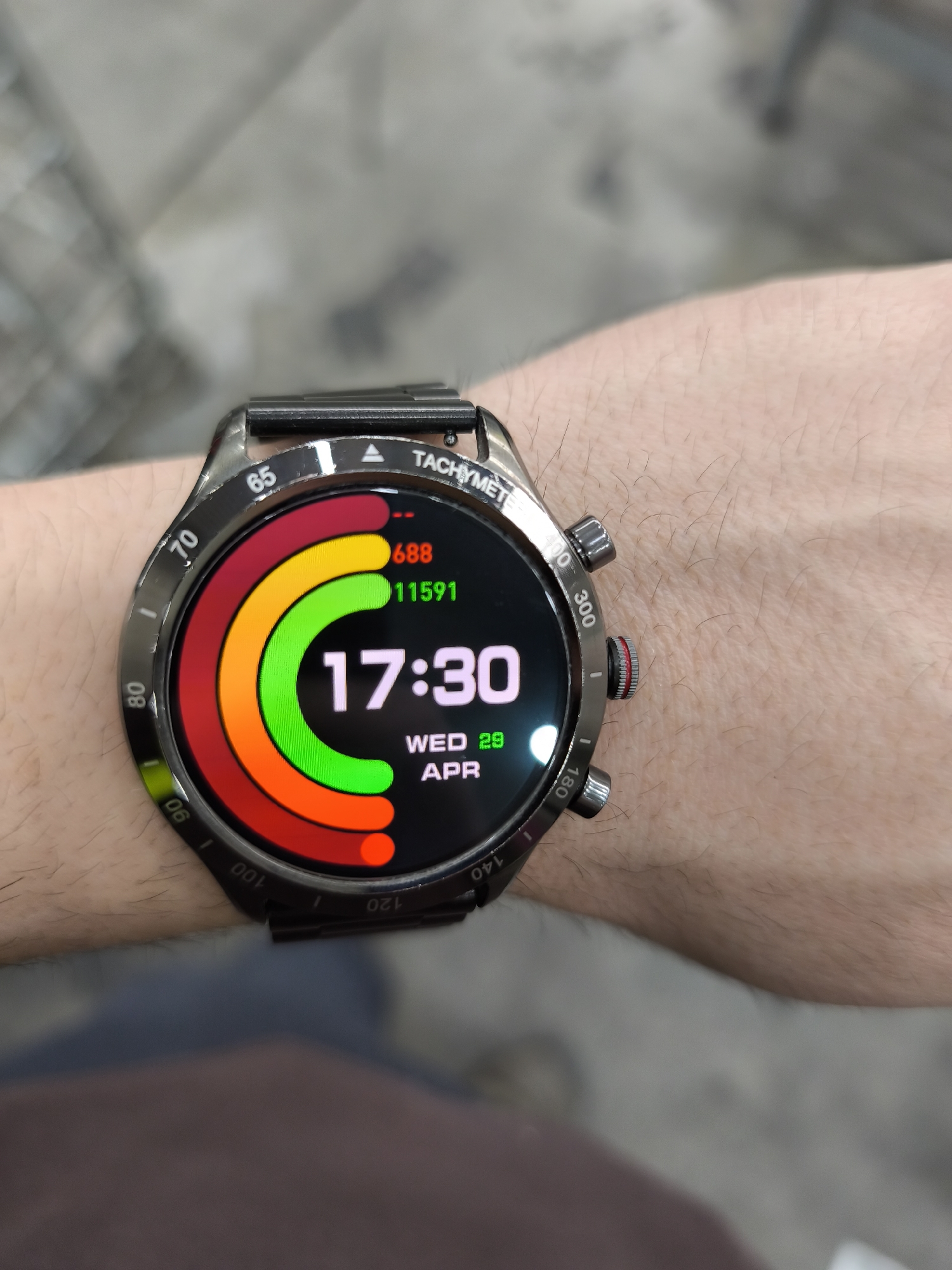A close-up shot captured in Burgas, Bulgaria, displays a person's left forearm and wrist, featuring a black smartwatch with a prominent round face and a metal link strap. The arm is fair-skinned and lightly hairy. The smartwatch has a dark, possibly gunmetal, finish on its bezel, which is inscribed with "TACHYMETER" and various numerical markings including 65, 70, 80, 90, 100, 120, 140, 180, 300, and 400. Three distinct chronometer-style pushers or crowns are visible on the right side of the watch case, one of which has a red band detail.
The smartwatch's digital display is active, showing the time as "17:30" (5:30 PM) in large white text. Below the time, the date "WED 29 APR" is displayed. The watch face also features three concentric, partially filled circular progress bars or rings: an outermost red ring, a middle orange ring, and an innermost green ring. Next to the red ring, the number "688" is visible, and adjacent to the green ring, the number "11591" is shown, likely indicating activity metrics such as calories burned and steps taken. The background is a blurred, light-colored indoor environment, providing no specific location cues beyond the context given.