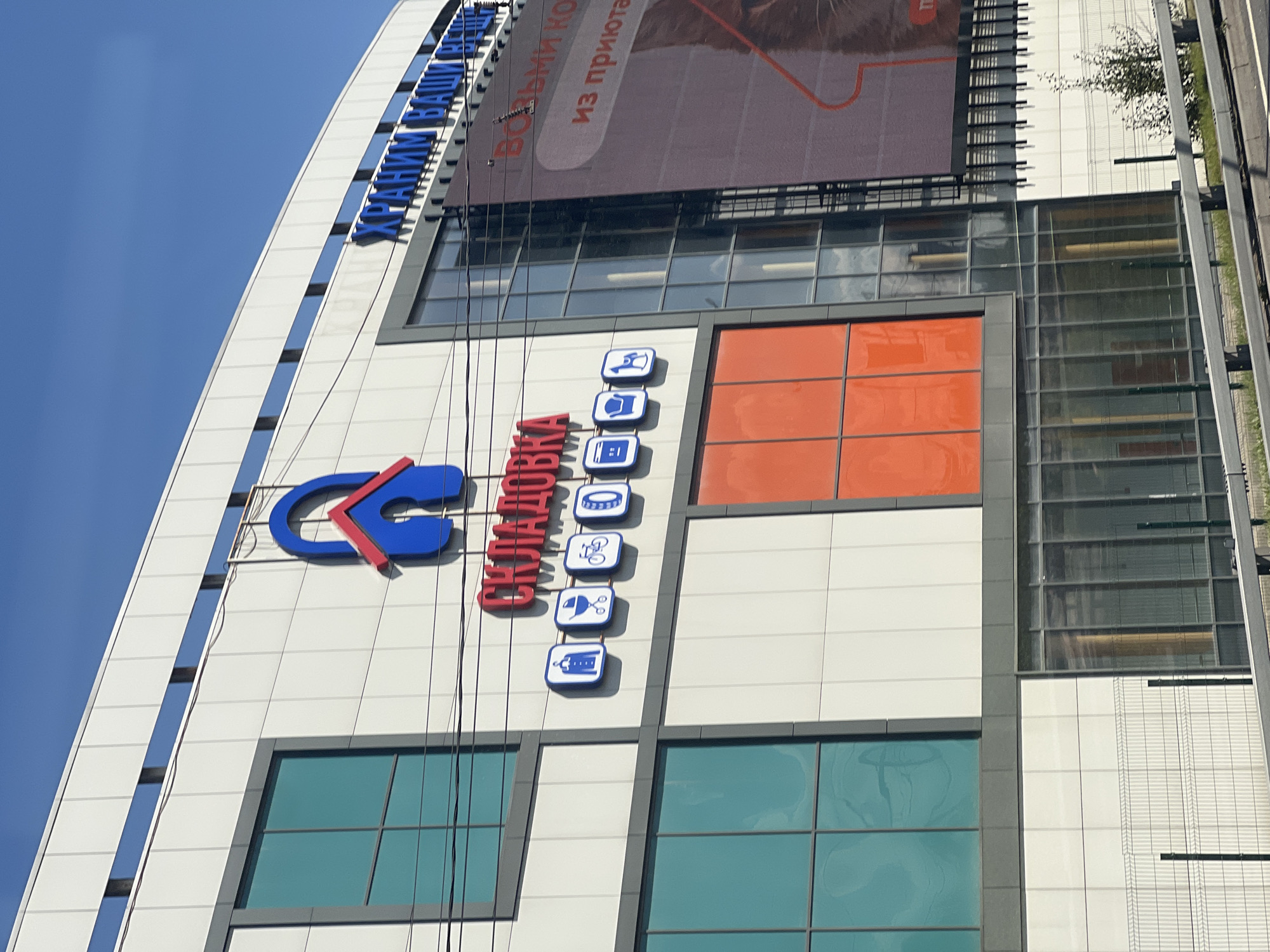 The image captures the exterior of a modern commercial building with a distinctive blue and red logo resembling a house within a lock. The building features large windows, some tinted turquoise and others appearing as opaque orange panels. Text in blue, Cyrillic lettering, reads "СКЛАДОВКА" (Skladovka) below the logo. To the right of this, another line of blue Cyrillic text reads "ХРАНИМ ВАШИ ВЕЩИ" (Khranim Vashi Veschi), meaning "We store your things." A large advertisement on the right side of the building features a pink background and some text, partially obscured. The building is situated in an urban environment, evidenced by the presence of power lines and a metal fence in the foreground, suggesting it is near a road. The sky above is clear blue. This building is located in the city of Khimki, Russia.