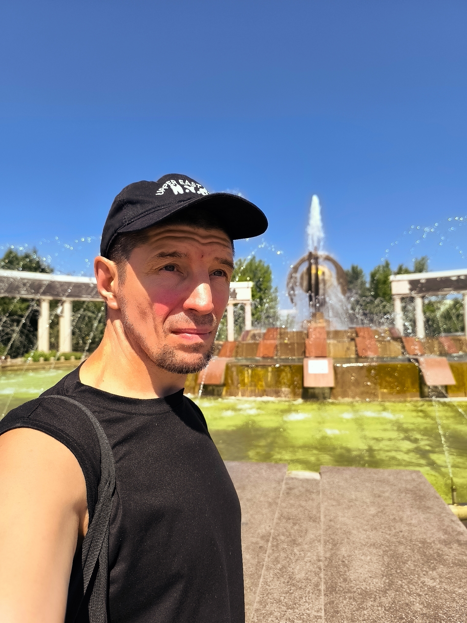 A man wearing a black t-shirt and a black baseball cap with white lettering is standing in front of a fountain in Qarağandı, Kazakhstan. The man is looking off to the right side of the frame. The fountain is large with water spraying high into the air. The water in the fountain's basin is a greenish color. In the background, there are trees and a blue sky.