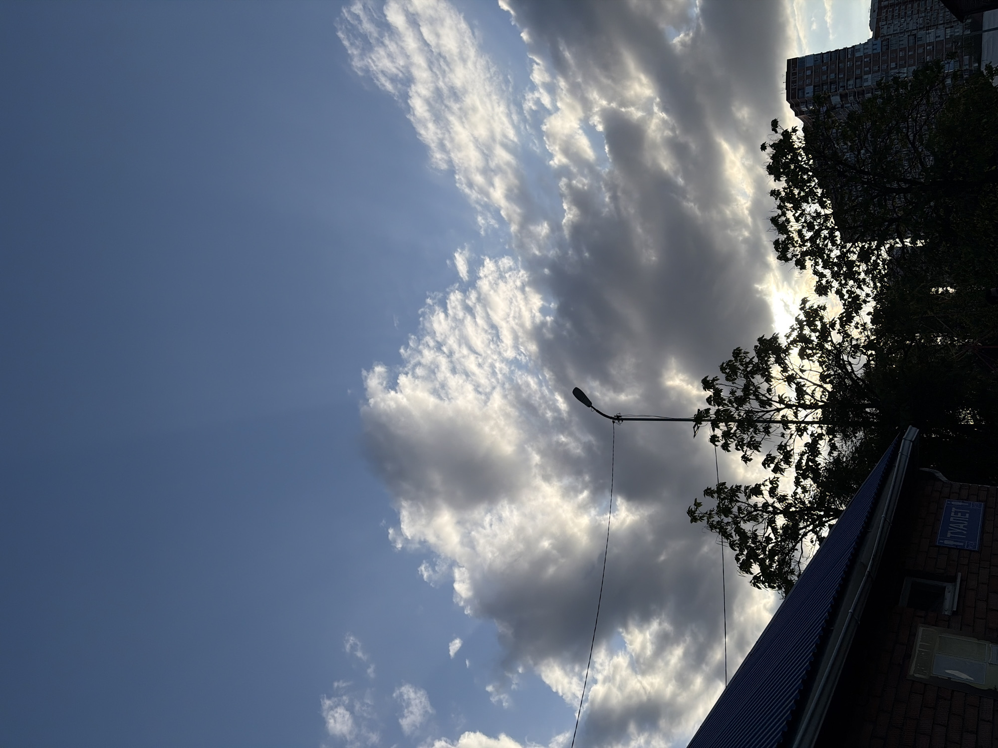The image shows a view of the sky with clouds, a building, a tree, and a street lamp. The building is a brick structure with a blue metal roof. There is a sign on the building that reads "ТУАЛЕТ" in Russian, indicating the presence of a public restroom. The tree is in the background, partially obscuring the building. The street lamp is positioned to the left of the building. The sky is filled with white and gray clouds, and there are patches of blue sky visible. Based on the sign and architectural style, the location is likely in a city, and considering the sign is in Russian, a city in Russia, possibly Krasnodar, is a plausible location.