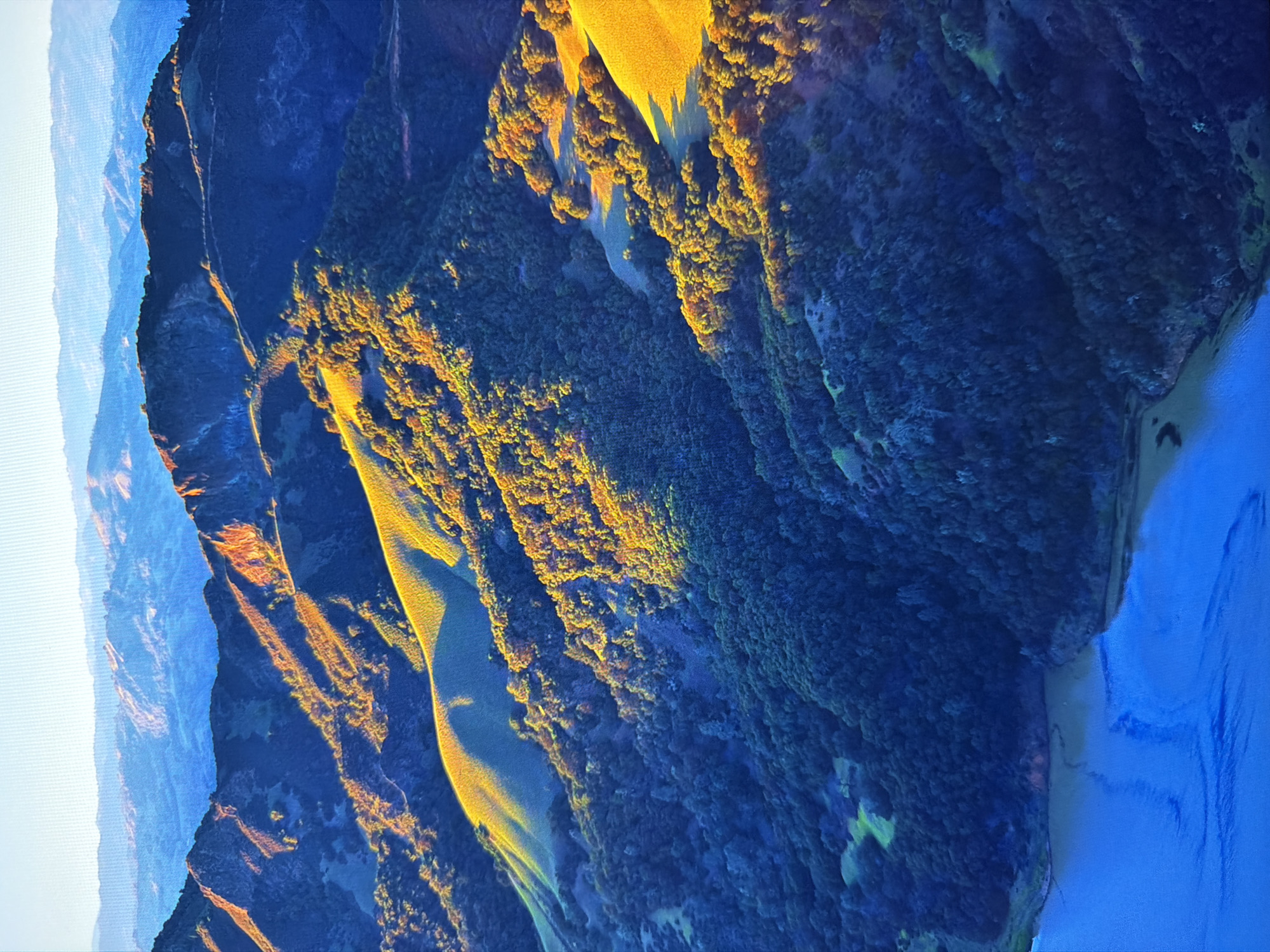 The image depicts a dramatic aerial view of rolling hills and mountains bathed in the golden light of sunrise or sunset. The terrain is rugged, with steep slopes covered in dense green vegetation, appearing as a dark, textured carpet in the shadows. Patches of grass or open ground on the hillsides are illuminated by the low-angle sun, creating striking contrasts of light and shadow. The distant mountain ranges fade into a hazy blue, suggesting a vast, expansive landscape. In the foreground, a body of water, likely a bay or inlet, reflects the sky and the surrounding hills. The water is a deep, vibrant blue with subtle ripples visible on its surface. The overall impression is one of natural grandeur and tranquility, with the interplay of light and shadow highlighting the contours of the land. The location is not Kyiv, Ukraine.