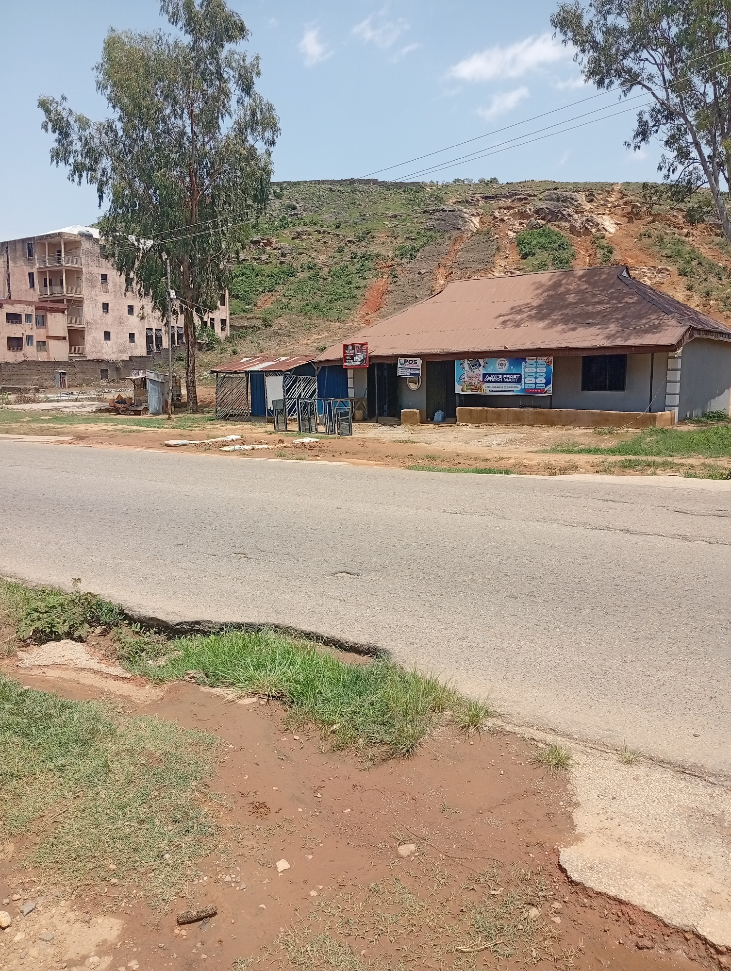 This image depicts a roadside scene in Gwong, Nigeria. A paved road occupies the foreground, with a grassy embankment to the left and a muddy, uneven terrain on the right. Beyond the road, a single-story building with a corrugated metal roof appears to be a small business, possibly a shop or mart, as indicated by a banner displaying "AJAY'S FROST & FRESH MART." Adjacent to this building are several smaller structures, some with tin roofs and others appearing to be open-air stalls displaying glass panes. In the background, a large, multi-story building with balconies and numerous windows stands partially obscured by a tall tree with sparse foliage. Behind the buildings, a rocky, vegetated hillside rises under a clear blue sky with scattered clouds. There are no people visible in the image. The overall impression is a somewhat undeveloped area with a mix of commercial and residential structures and natural landscape.
