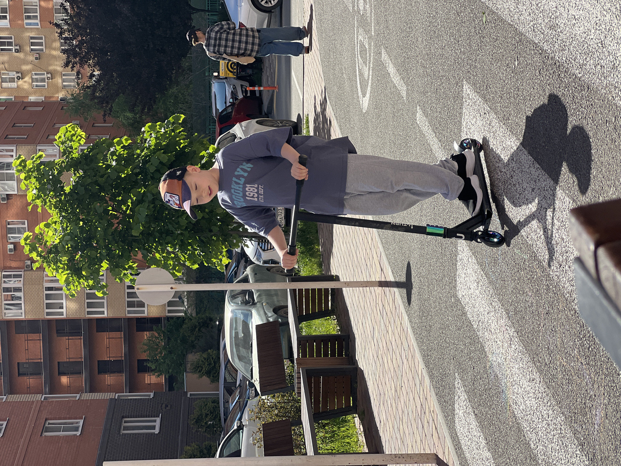 A boy rides a scooter across a pedestrian crosswalk in Krasnodar, Russia. The boy wears a baseball cap, t-shirt, and sweatpants. A man stands in the background on the sidewalk. Buildings, cars, trees, benches, and a street sign surround the scene.