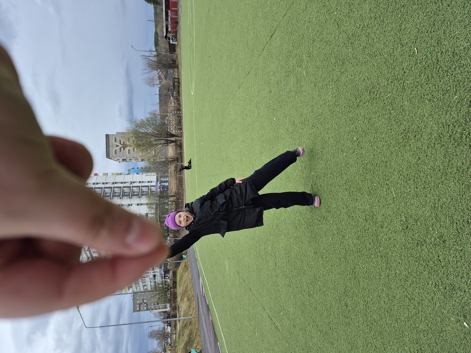 An outdoor, rotated full shot captures a person enthusiastically posing on an artificial green turf sports field in Chaikovsky, Russia, under an overcast sky. The image is oriented vertically with the horizon running down the left side. The main subject, a child or young adult, is centered and wearing a black jacket, black pants, and a purple beanie. They are smiling broadly with an open mouth, facing the camera, and have their arms outstretched and one leg lifted, suggesting a playful or energetic action.
In the immediate foreground, a blurred human hand extends from the left, partially obscuring the view. The green artificial turf of the field is clearly visible, with white lines indicating a sports pitch, possibly for soccer.
The background reveals an urban or suburban setting. Multi-story apartment buildings, some with colorful facades, line the horizon. Bare trees and utility poles are scattered across the midground. A faint road or path runs alongside the field. The overall atmosphere suggests a cool, possibly early spring or late autumn day, with no strong sunlight. No discernible text is visible in the frame.