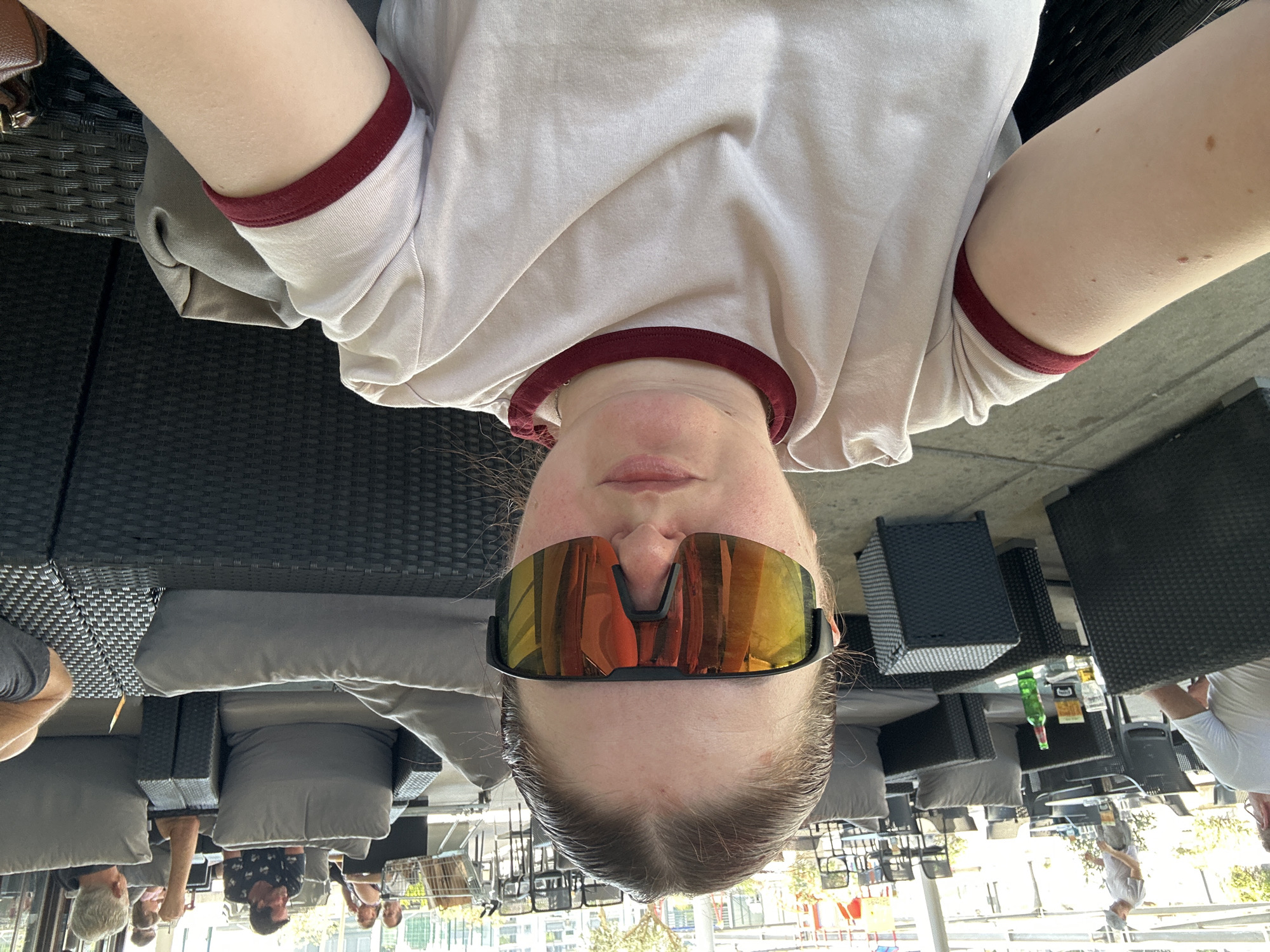 The image shows a young woman taking a selfie in an outdoor seating area, possibly a cafe or restaurant. She is wearing a light-colored t-shirt with burgundy trim on the collar and sleeves, and large reflective sunglasses. Her hair is pulled back. The background reveals a seating area with dark wicker furniture, grey cushions, and tables. Several other people are visible in the background, some seated at tables and others walking. A green bottle and glasses are on a table in the middle ground. The environment appears to be a commercial outdoor space.
The image does not contain any information that can be used to determine the specific location within the city of Rijeka, Croatia.
